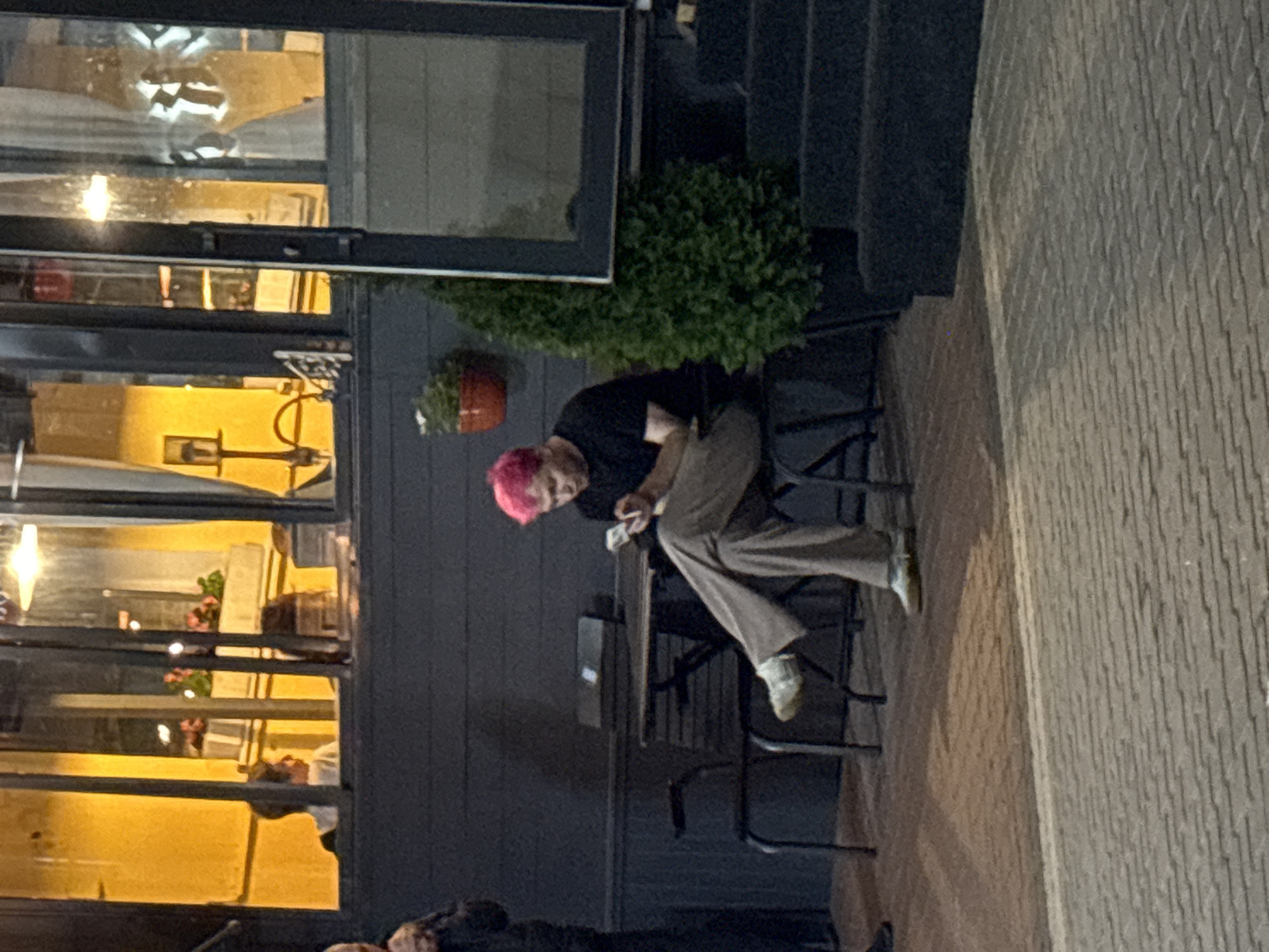 The image shows a person with pink hair sitting outdoors at a table with chairs, looking at their phone. The person is wearing a black t-shirt and light-colored pants. The setting appears to be an outdoor seating area of a restaurant or cafe, with a building in the background. Inside the building, seen through windows, are indications of artificial lighting and plants. A potted plant and a small evergreen tree are visible outside. The ground is paved. The image does not provide any specific landmarks or visual cues to definitively place it within Vitsebsk, Belarus.