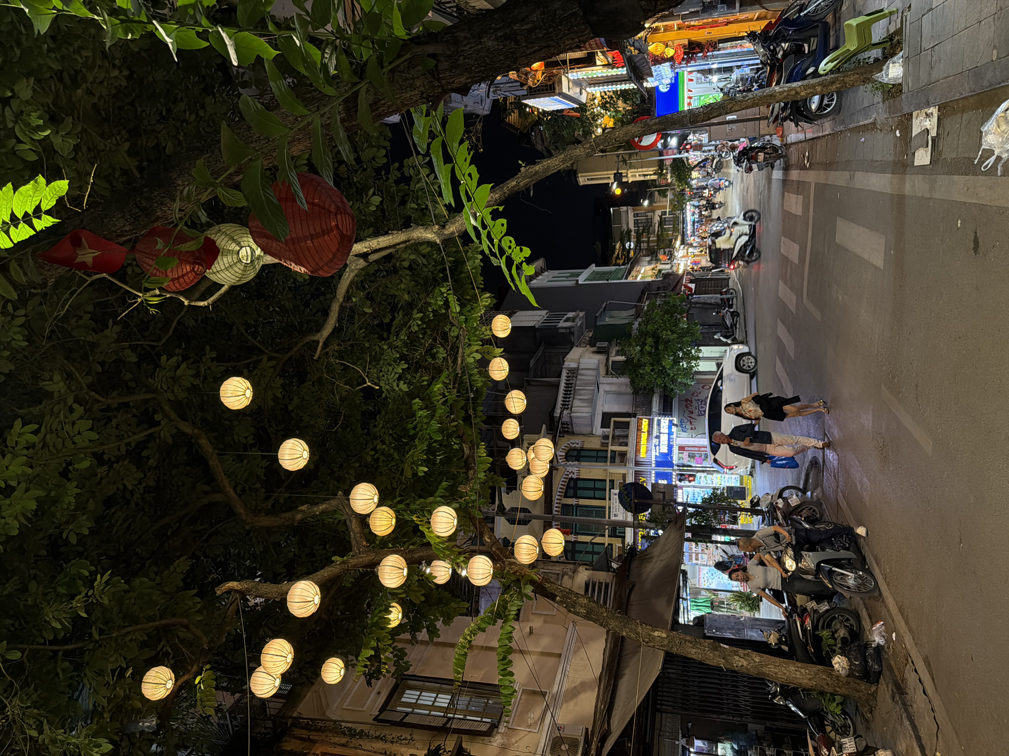 This is a night scene depicting a lively street in Hà Nội, Vietnam. The image is captured from an elevated perspective looking down the street.
On the left side, large trees with dense, dark green foliage line the street, forming a natural canopy. Many decorative lanterns are strung between the branches. These include several red, pleated, cylindrical lanterns, and one prominent red lantern featuring a yellow star, reminiscent of the Vietnamese flag. Below these, numerous spherical white or cream-colored lanterns, with a ribbed texture, are suspended, casting a warm glow.
On the right side of the street, multi-story buildings house various businesses, their brightly lit signs illuminating the sidewalk. The architecture suggests a typical urban setting. A red circular sign with a diagonal line, indicating "No Parking," is visible near the top right of the street.
The street itself is asphalt with white striped road markings. Numerous motorbikes are parked haphazardly along the sidewalks and on the street, a common sight in Vietnamese cities. Several people are visible: two individuals, a man and a woman, walk side-by-side on the right sidewalk, and further down, two men stand beside parked motorbikes, seemingly engaged in conversation or waiting. Other figures are visible in the distance, suggesting a bustling environment.
The overall atmosphere is vibrant and energetic, typical of an evening in a Southeast Asian city. The glowing lanterns add a festive or charming touch to the urban landscape. The time of day is clearly night, with the dark sky contrasting with the artificial lights from shops and lanterns.