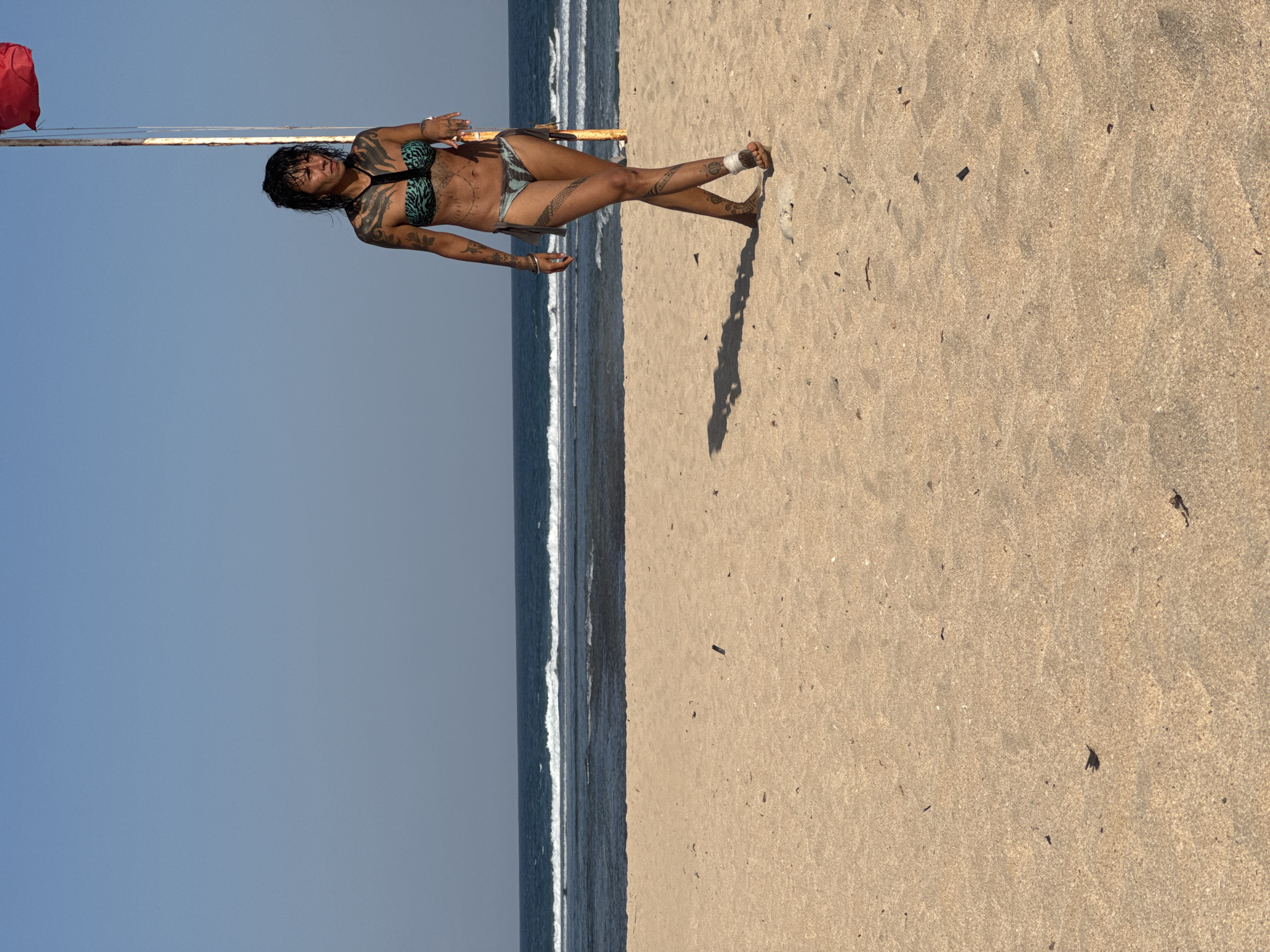 A tattooed woman in a bikini walks on a sandy beach in Kuta Selatan, Indonesia. She has dark, wet hair and several intricate tattoos covering her arms, torso, and legs. She is wearing a patterned bikini top and bottoms. A red flag flies on a pole behind her, and the ocean with gentle waves is visible in the background under a clear blue sky. The sand is light brown and scattered with small debris. The woman's shadow stretches out before her on the sand.