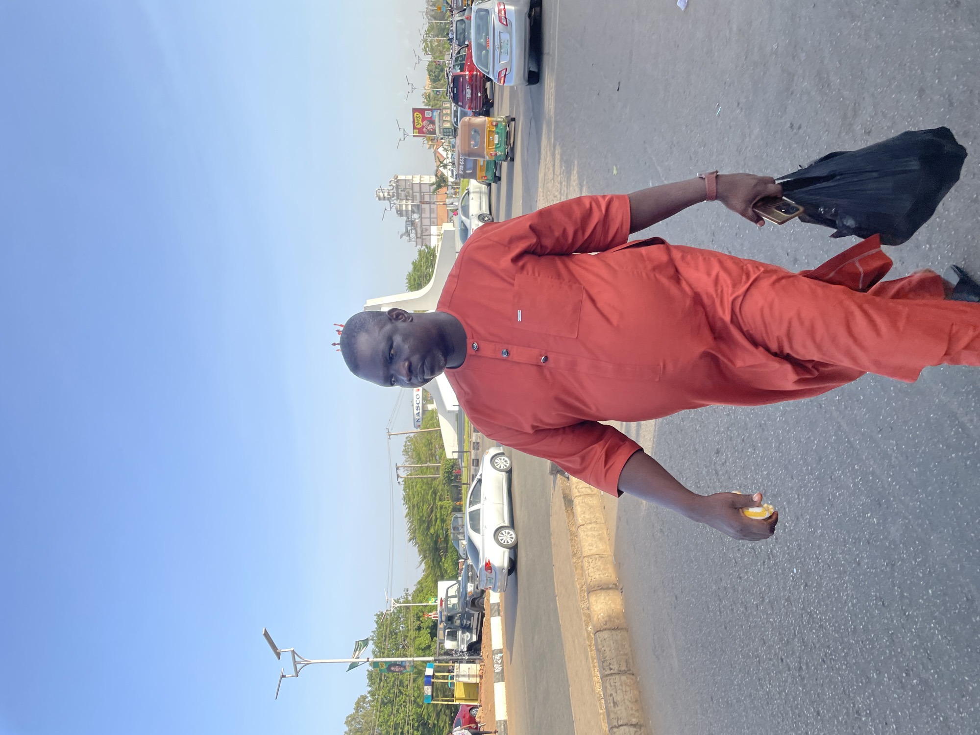 A medium-angle full shot depicts a dark-skinned man wearing an orange-red tunic with short sleeves and dark trousers, standing on an asphalt road. The man is carrying a black bag in his left hand, which also holds a smartphone. In his right hand, he is holding an object with yellow and white elements.
Behind the man, the scene reveals an urban environment. The background includes a number of vehicles, including cars and a green and yellow auto rickshaw, indicating street traffic. The sky is blue, suggesting a sunny day. Streetlights and trees line the road. In the middle distance, there are buildings, including one with a sign that reads "NASCO," and a structure that looks like an architectural monument. The scene is likely taking place on a street in Jos, Nigeria.