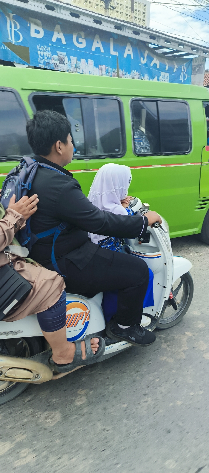 A green bus with the words “BAGALA JAY” in blue letters on the side is parked on a road in Sukatani, Indonesia (latitude: -6.611, longitude: 107.411). A white motorbike is stopped in front of the bus, with a young man and a young woman on board. The man is wearing a black jacket and black pants and is holding the handlebars of the motorbike. The woman is wearing a white hijab and is holding onto the man. A man with brown pants, a black shirt, and a blue backpack is sitting behind the woman. The woman is holding on to the man in front of her, and her hand is visible in front of the other man’s face. The three are riding a white motorbike with a blue and orange sticker on it. The motorbike has a large, grey exhaust pipe. The road surface is grey asphalt.