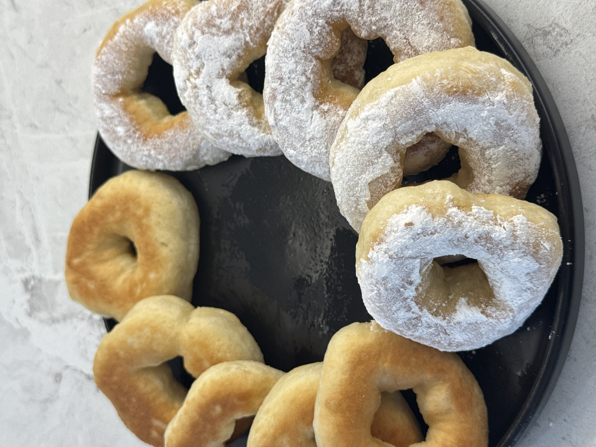 A close-up, overhead shot shows a black plate filled with donuts. The donuts are arranged in a circle, with some dusted with powdered sugar and others plain. The plate sits on a light-colored, textured surface. The image does not contain any information about the city of Lyubertsy, Russia.