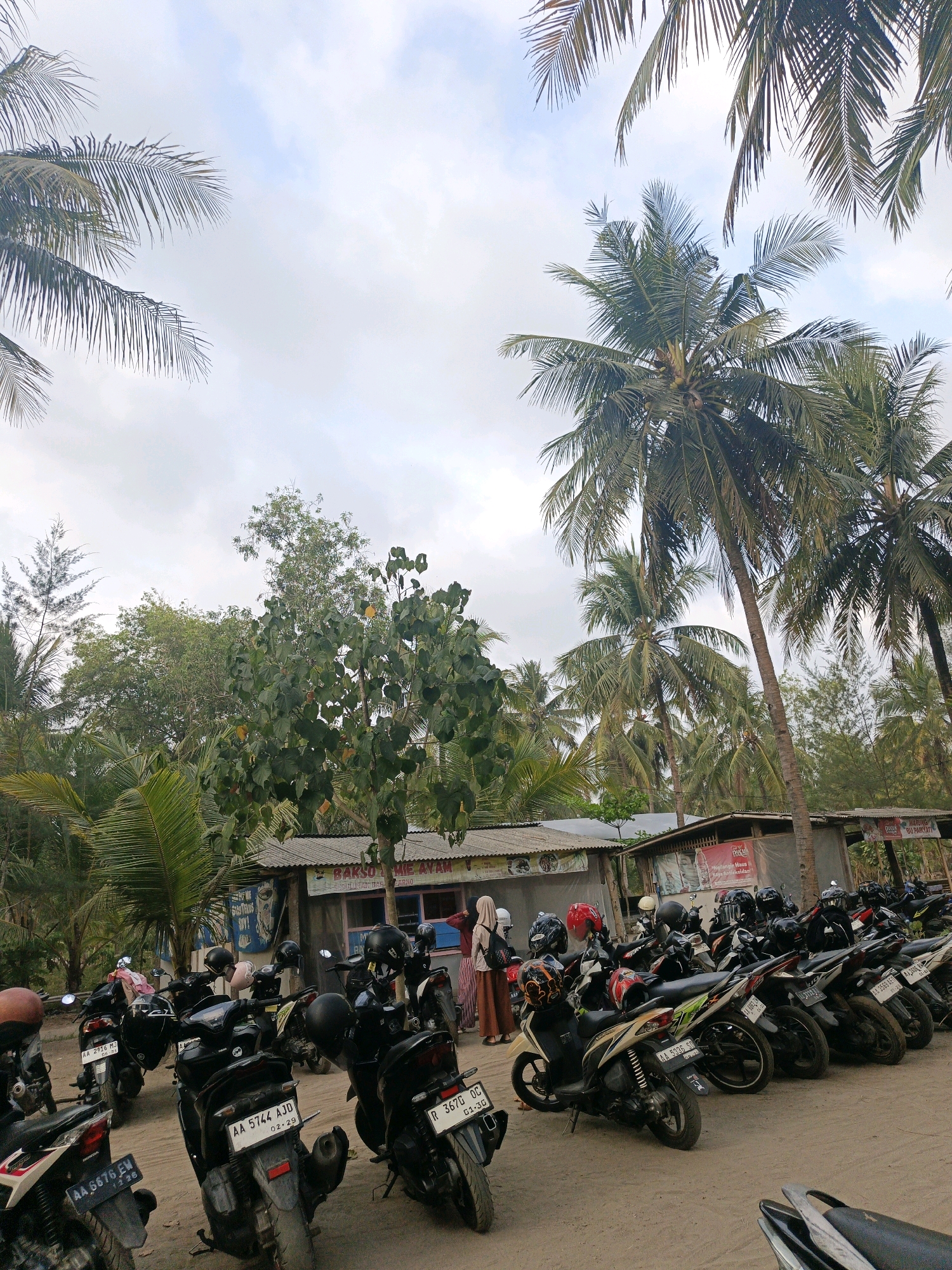 The image depicts a parking area for motorcycles in Tambakmulyo, Indonesia, likely near a beach or coastal area. Several motorcycles are parked in rows on a dirt or sand surface. In the background, there are simple, open-sided buildings with corrugated metal roofs, suggesting food stalls or small shops. One building has a sign that reads "BAKSO MIE AYAM," indicating it sells noodle soup and chicken meatballs. Palm trees, characteristic of tropical coastal regions, are abundant, with their fronds reaching towards a cloudy sky. Two individuals are visible near the buildings, one wearing a headscarf. The overall atmosphere is casual and suggests a destination popular with motorcycle-riding visitors.