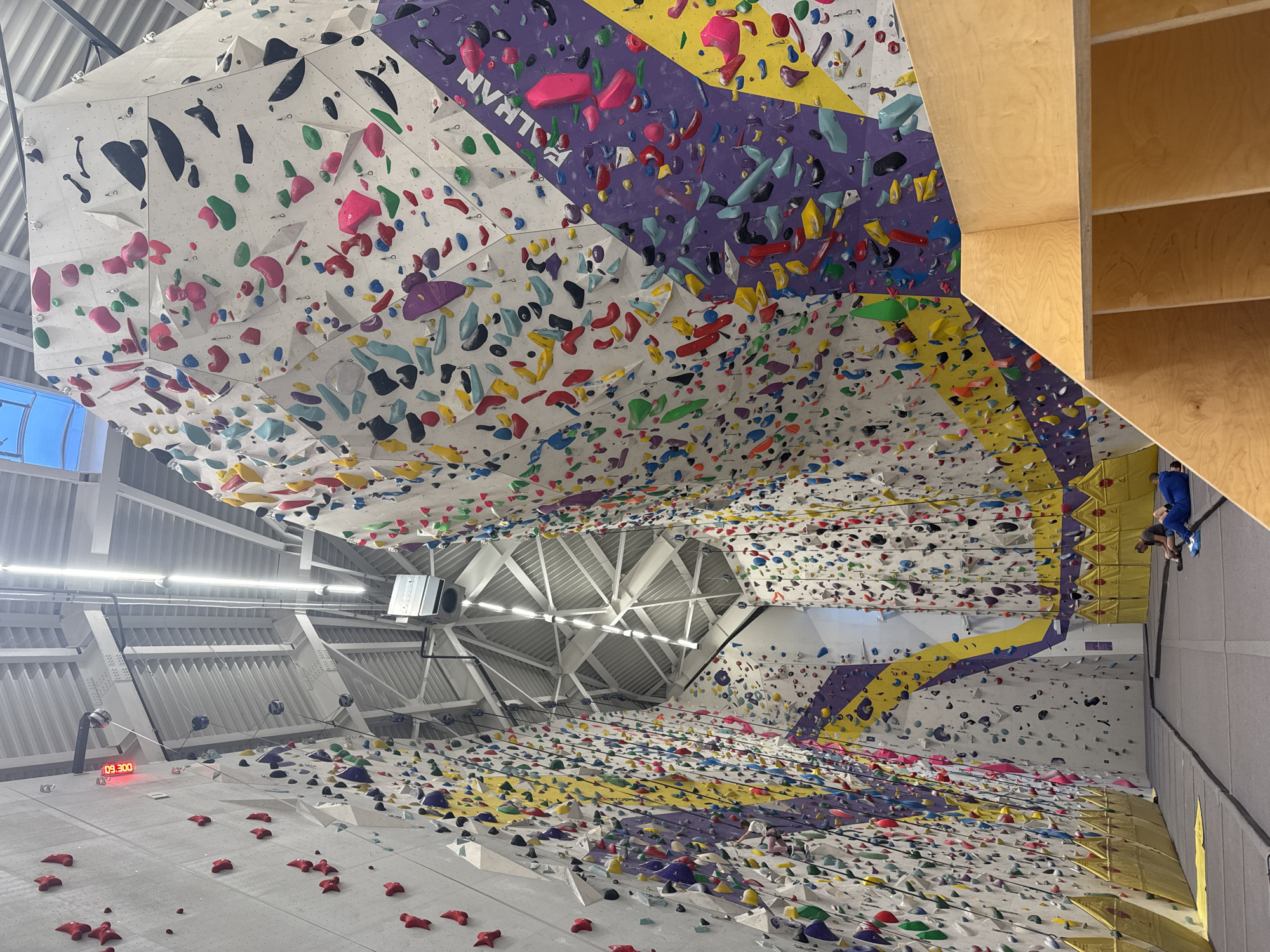 The image depicts an indoor rock climbing gym. The climbing walls are covered with numerous colorful holds. The walls are angled and extend towards the ceiling, creating an overhang. The ceiling structure is visible, featuring metal beams and lighting fixtures. A digital clock displays 09:30:00. To the right, two individuals are climbing. Based on the name "KLIMATRON" visible on the wall, the location is likely the Klimatron climbing gym in Sofia, Bulgaria.