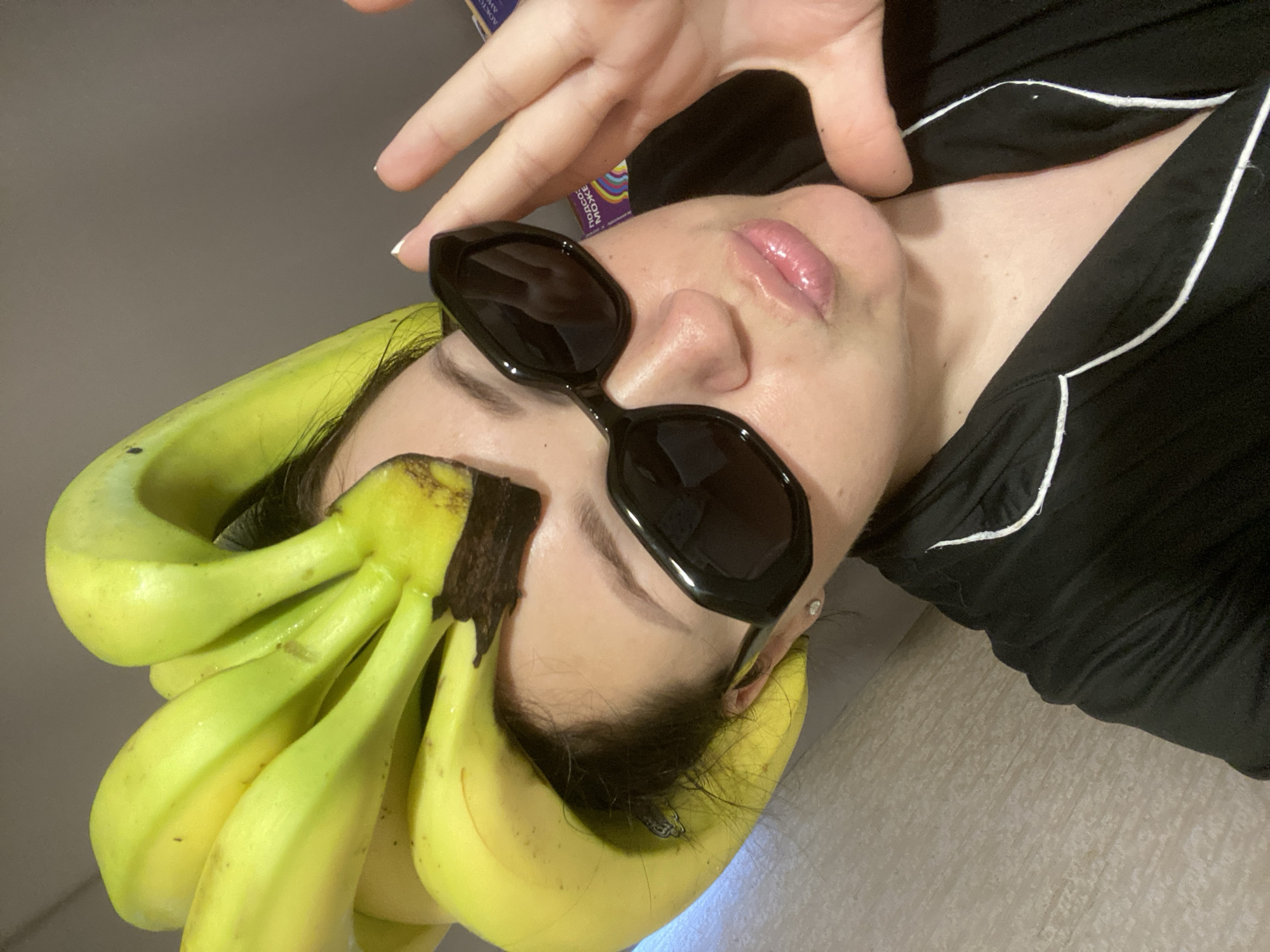 A person, presumably female, is lying down with a bunch of bananas arranged on their head like a hat. The person is wearing black sunglasses and a black shirt with white piping. Their hand is raised slightly above their head. The background is a neutral gray color. There is a glimpse of a product box in the upper left corner of the image. There is no definitive indication that the location is Sochi, Russia.