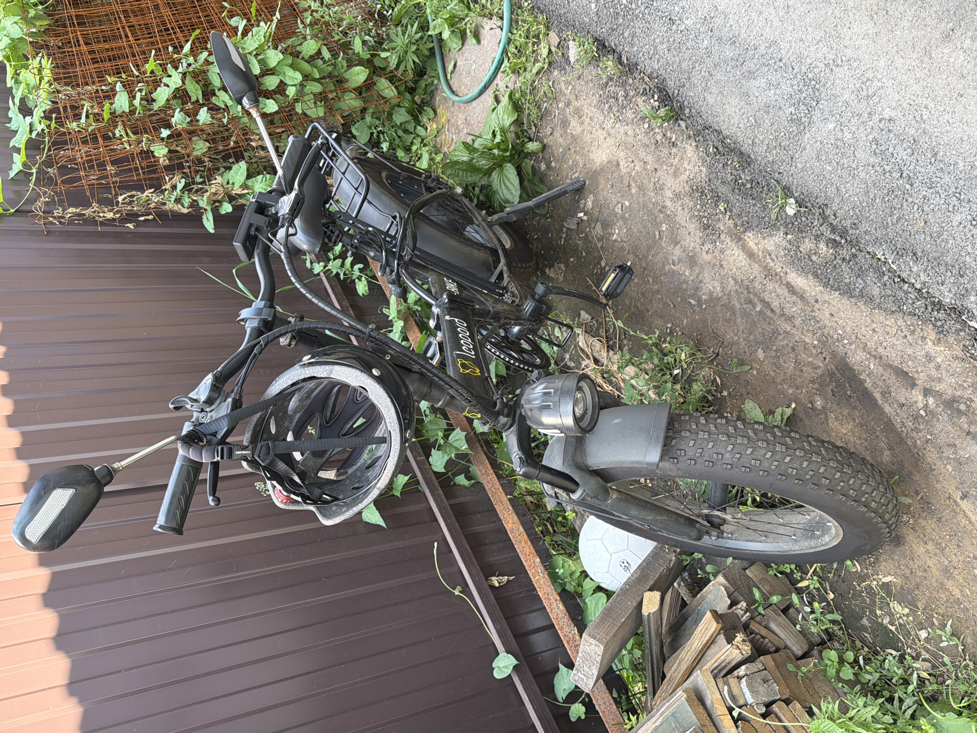 The image depicts a black electric bicycle parked next to a brown corrugated metal fence. A grey and black helmet is hanging from the handlebars of the bicycle. In the background, there is overgrown green vegetation climbing on a rusty metal grid. To the left of the bicycle, a pile of wooden planks and a white soccer ball are visible. The ground is a mix of dirt and asphalt. The scene appears to be in an outdoor urban or suburban setting, possibly a backyard or alleyway in the city of Orenburg, Russia.