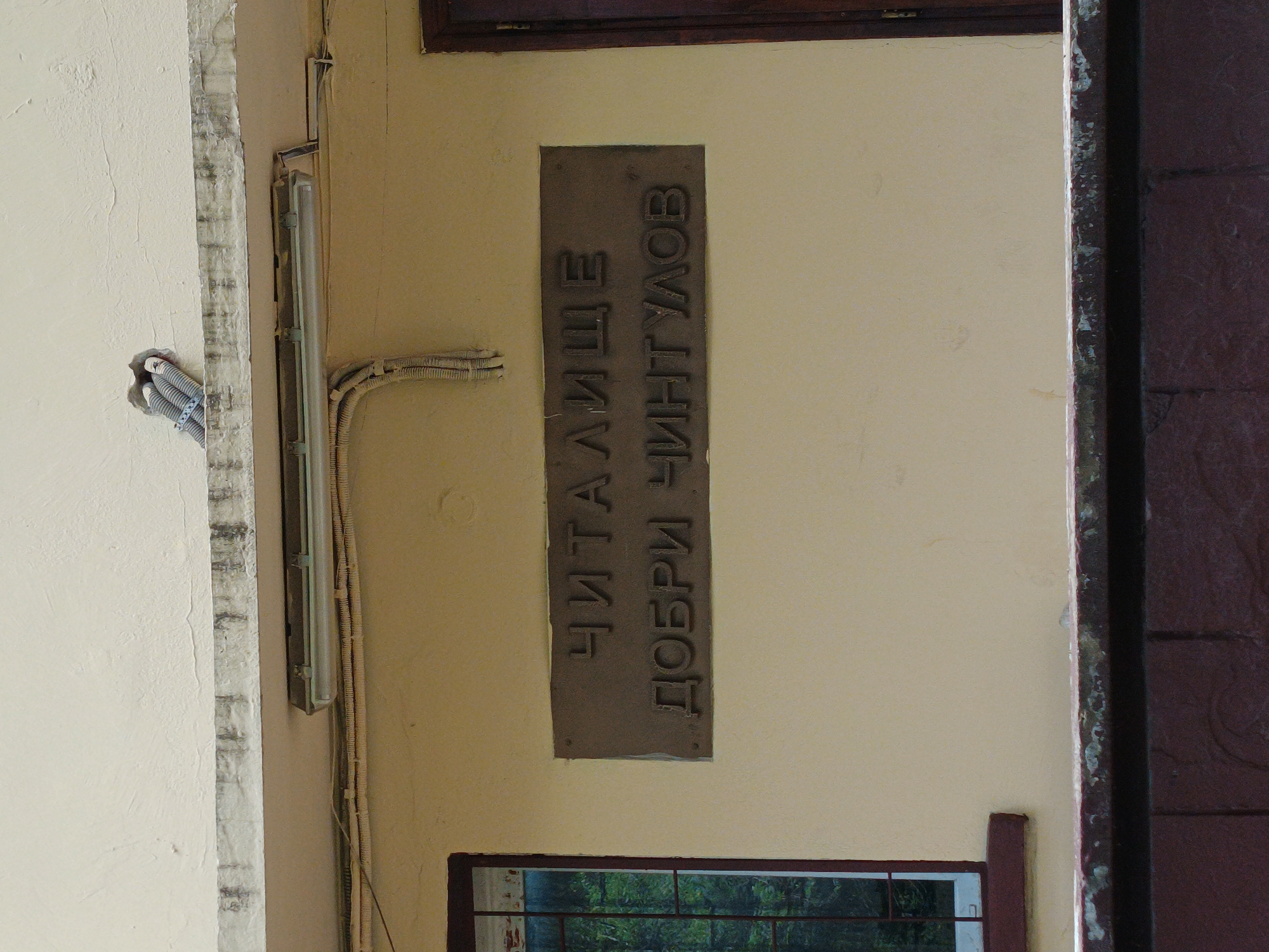 A close-up, slightly rotated image shows a section of a building facade in Sofia, Bulgaria. A beige wall is visible, featuring a metal plaque with Cyrillic lettering that reads "ЧИТАЛИЩЕ ДОБРИ ЧИНТУЛОВ" (Chitalishte Dobri Chintulov). Below the plaque is a window with a dark brown frame. The view through the window is obscured, but greenery is visible. A fluorescent light fixture is mounted horizontally on the wall, with electrical conduits running alongside it. To the left, electrical wires are seen emerging from the wall. A section of a dark brown door frame is visible on the right side of the image.