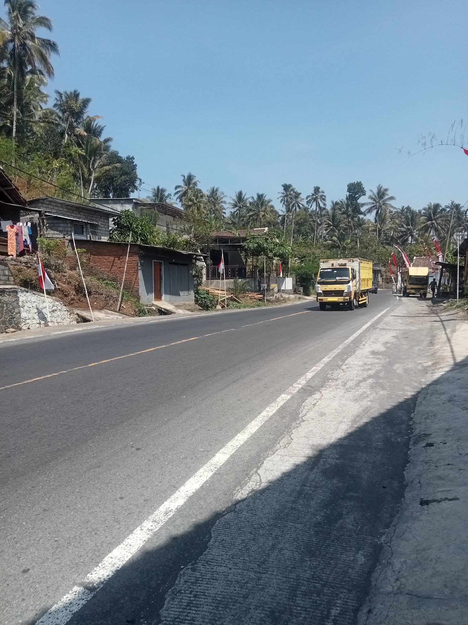 The image depicts a street scene in Tlogosari, Indonesia, on a clear, sunny day. A two-lane asphalt road runs through the middle of the frame, with a white line dividing the lanes and a yellow line on the outer edge of the right lane. The left side of the road features a slightly elevated embankment with several modest structures, including a brick building with clothes hanging outside and an adjacent building with a gray facade and a wooden door. A stone wall borders the embankment. Several Indonesian flags are visible, one red and white on the left embankment and two more further down the road.
On the right side of the road, there is a paved shoulder and sidewalk area. A yellow truck with a white cargo box is driving away from the viewer down the road. Further in the distance, another yellow truck is parked, with a person standing next to it. A line of palm trees and other vegetation can be seen on either side of the road, extending up the hillsides in the background, under a clear blue sky. The overall impression is a rural or semi-rural setting in Indonesia.