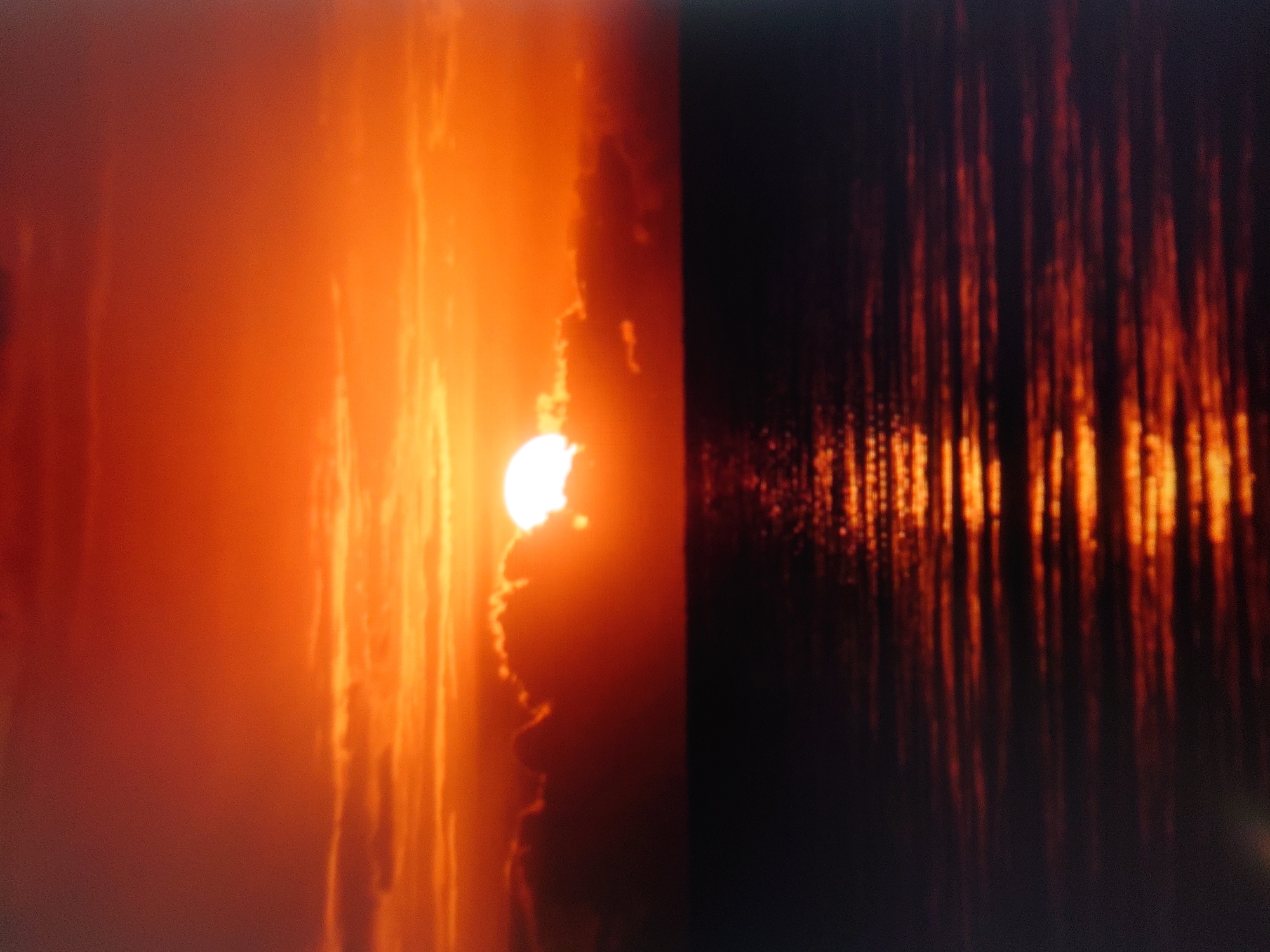 A hazy, vibrant sunset casts an intense orange and red glow across the sky and the still ocean. The sun, a bright white disc partially obscured by dark clouds, is low on the horizon, creating a dramatic silhouette against the fiery sky. The water reflects the vibrant colors of the sunset, with streaks of orange and yellow shimmering on its dark surface. The scene is serene and evokes a sense of calm, despite the intense colors. There are no people or specific landmarks visible to indicate a precise location within Belorechensk, Russia.