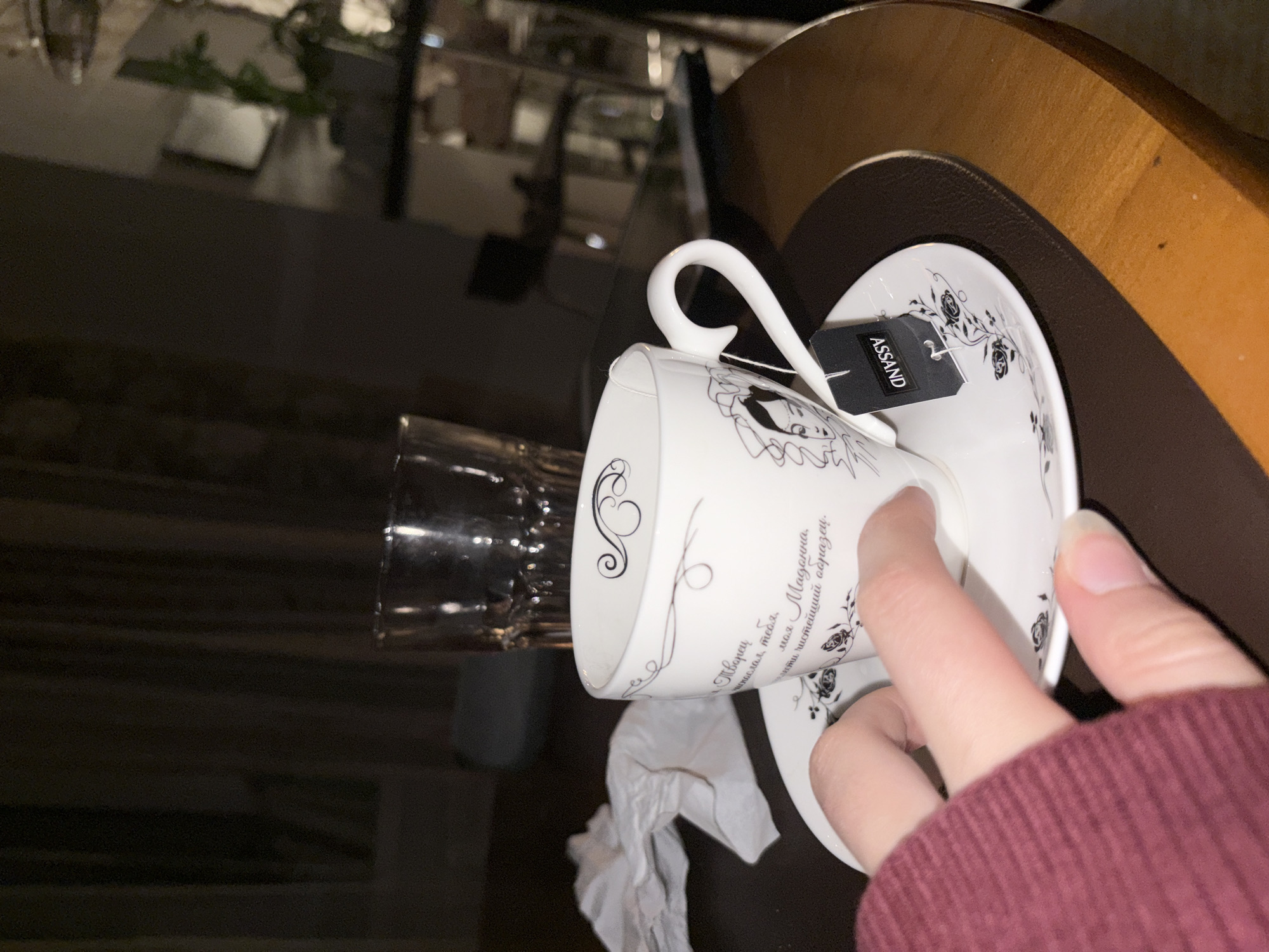 The image shows a close-up view of a hand reaching for a white teacup with black filigree and a portrait on it, resting on a saucer. A black tea bag tag with the word "ASSAND" is visible. A crumpled white napkin is to the left of the cup, and a glass is in the background. The scene is set on a wooden table, with a dark, out-of-focus background suggesting an indoor environment, possibly a cafe or home. There are no identifiable landmarks or clues to pinpoint the location to Chelyabinsk, Russia.