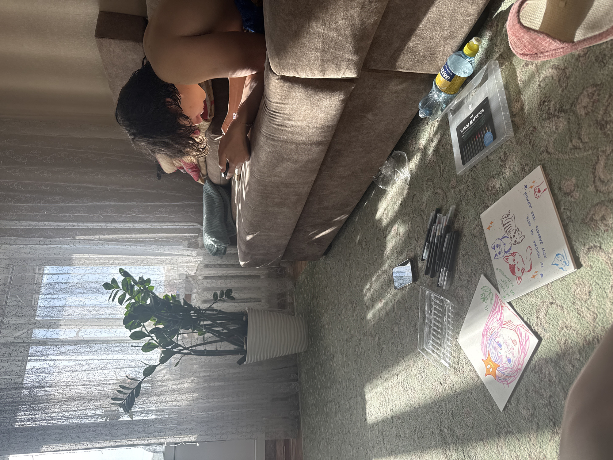 A room interior in Uryupinsk, Russia, is depicted with a person lying on a brown sofa. The person has dark, curly hair and is wearing blue shorts. On the floor, which is covered with a patterned rug, are drawing materials: markers, drawings of cats and a character with pink hair, a water bottle labeled "Active", and a case for markers. A ZZ plant in a white pot is positioned in front of a window covered with blinds. A pink slipper is partially visible on the rug. The scene is illuminated by sunlight.