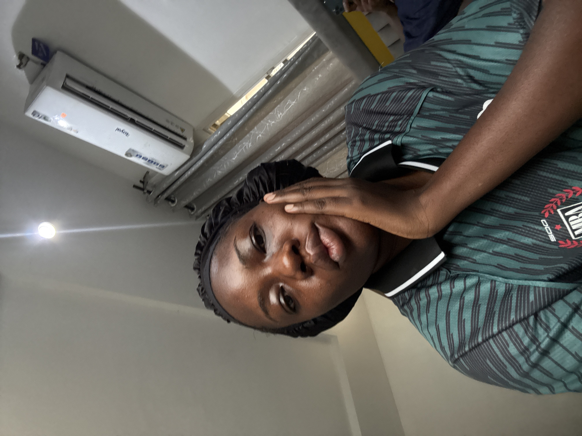 A person wearing a black head covering and a green and black patterned shirt is looking at the camera with their hand on their face. The setting appears to be indoors. An air conditioning unit is visible on the wall, and a curtained window is nearby. There's a light fixture on the ceiling. The location is Iddo Tudunwada, Nigeria.