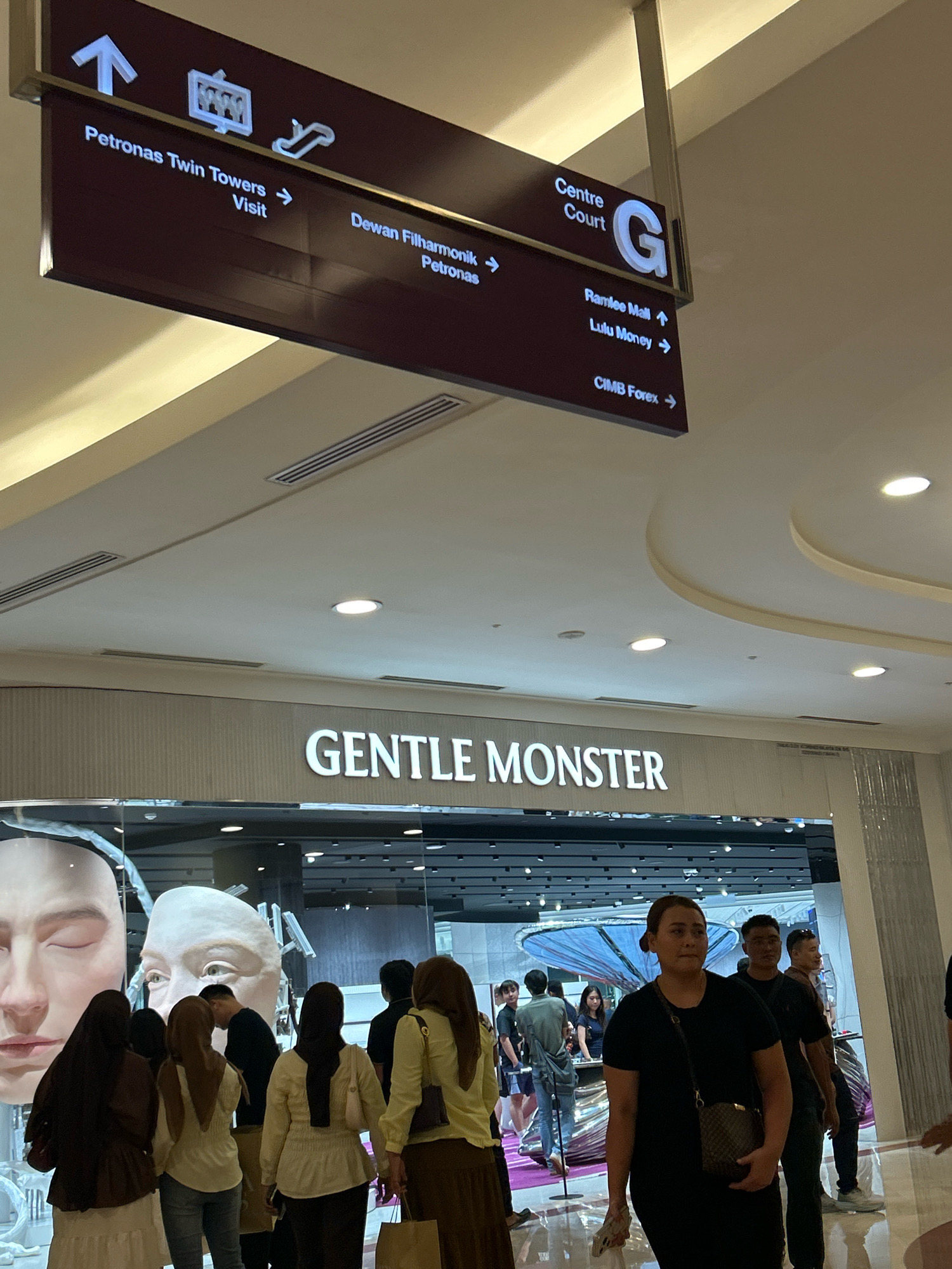 The image depicts a signpost in a public indoor space, likely a shopping mall in Kuala Lumpur, Malaysia. The signpost is dark brown with illuminated white text and directional arrows. It indicates directions to "Petronas Twin Towers Visit", "Dewan Filharmonik Petronas", "Centre Court G", "Ramlee Mall", "Lulu Money", and "CIMB Forex". Below the signpost, the storefront of "GENTLE MONSTER" is visible, an optical shop featuring large, artful displays of faces. Several individuals, predominantly women wearing hijabs, are seen browsing in front of the store. The environment appears well-lit and modern, with reflective floors and suspended ceiling elements.