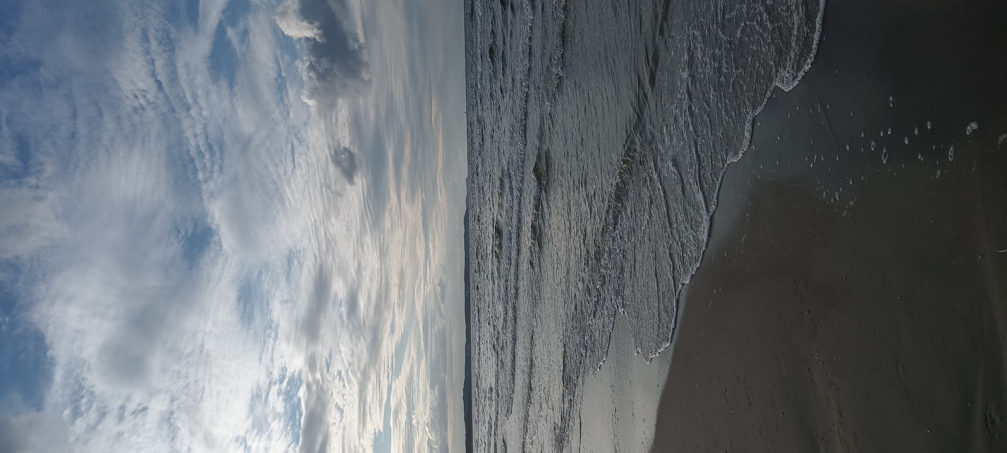 This is a vertical, full-frame, eye-level shot of the Baltic Sea coastline in Świnoujście, Poland. The image captures a cloudy sky above a calm sea with gentle waves lapping onto a dark, wet sand beach. The foreground is dominated by the beach, showing subtle footprints and the glistening, dark sand where the water has receded. The sea occupies the midground, with distinct lines of incoming waves creating a textured surface. A thin strip of land, possibly a distant shore or dunes, is visible on the horizon under a sky filled with various types of clouds, ranging from wispy cirrus to more substantial cumulonimbus formations. The overall lighting suggests it is either early morning or late afternoon, with diffused sunlight filtering through the clouds, creating a subdued and atmospheric scene. No people or man-made objects are clearly visible.