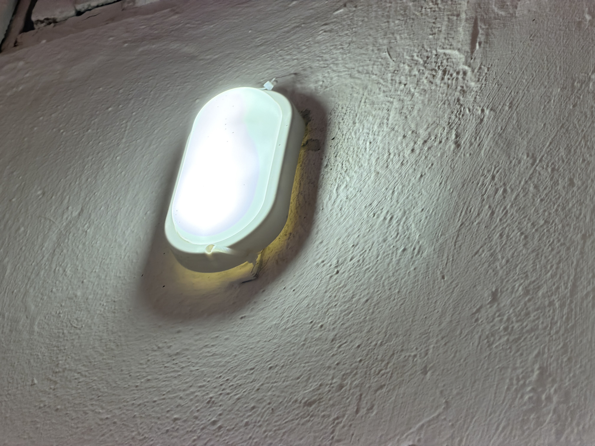 An oval-shaped, white light fixture is mounted on a textured, light-colored wall in Okha, Russia. The light emitted from the fixture is bright and appears to be a cool white. The wall has a rough, uneven surface with visible brush strokes or trowel marks. A shadow is cast by the light fixture onto the wall, creating a subtle contrast. The image focuses on the light and the texture of the wall, with no other objects or people visible. The scene appears to be indoors, possibly in a utility area or hallway.