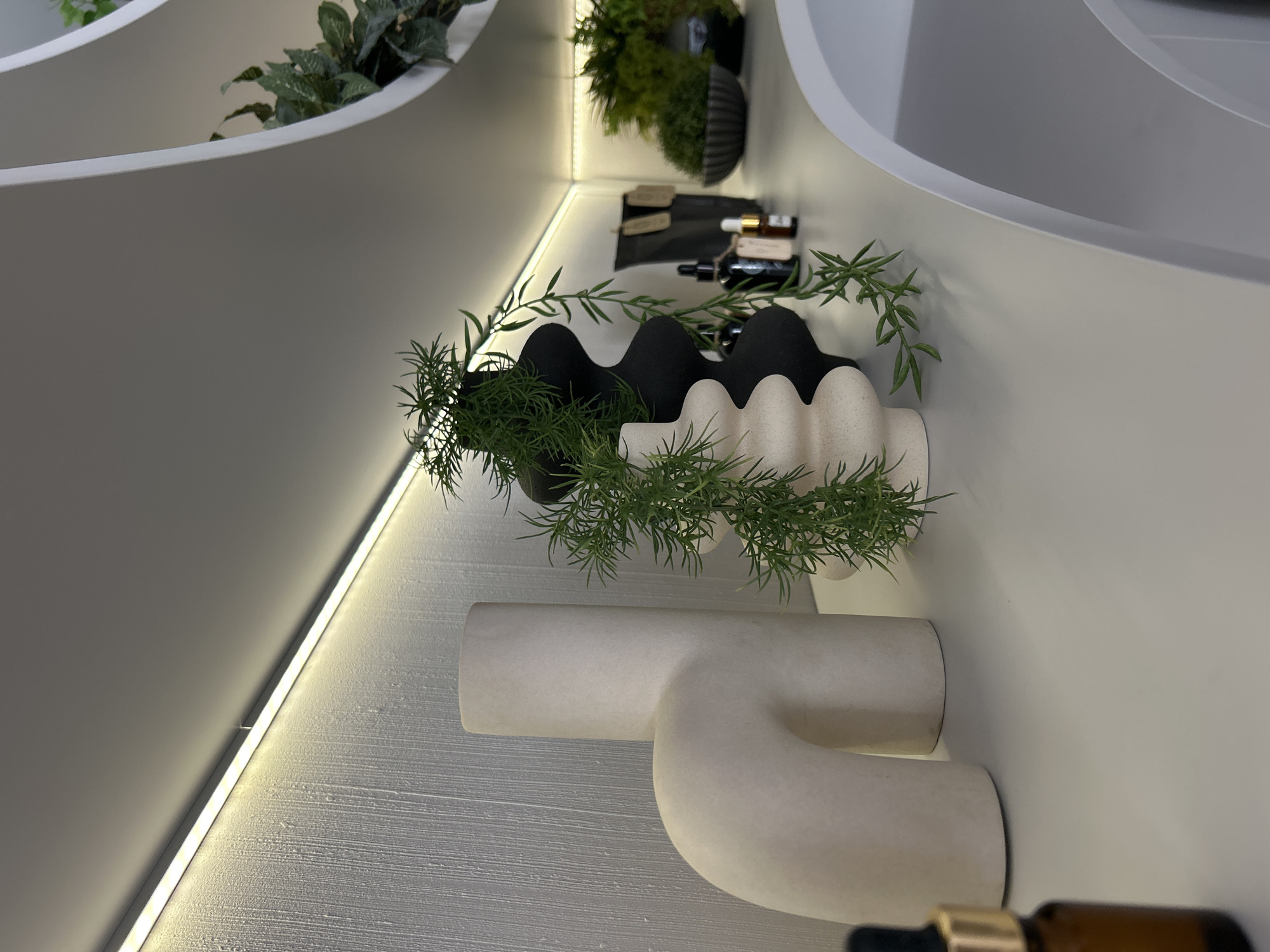 The image shows a close-up view of a shelf with various decorative items. On the left, there is a large, curved, light-colored vase. To its right are two more vases, one black and one cream-colored, with a cascading green plant draped over them. Further to the right, there are two dark bottles with tags, and behind them, a dark bowl filled with a lush green plant. The shelf itself is a light color, and a strip of warm LED lighting runs along the top of the wall behind it. The background wall has a textured finish. The scene appears to be an interior, possibly a home or retail display. There are no people or animals visible in the image. The overall aesthetic is modern and minimalist, with a focus on organic shapes and natural elements. The specific location of Votkinsk, Russia, cannot be determined from the visual information provided in the image.