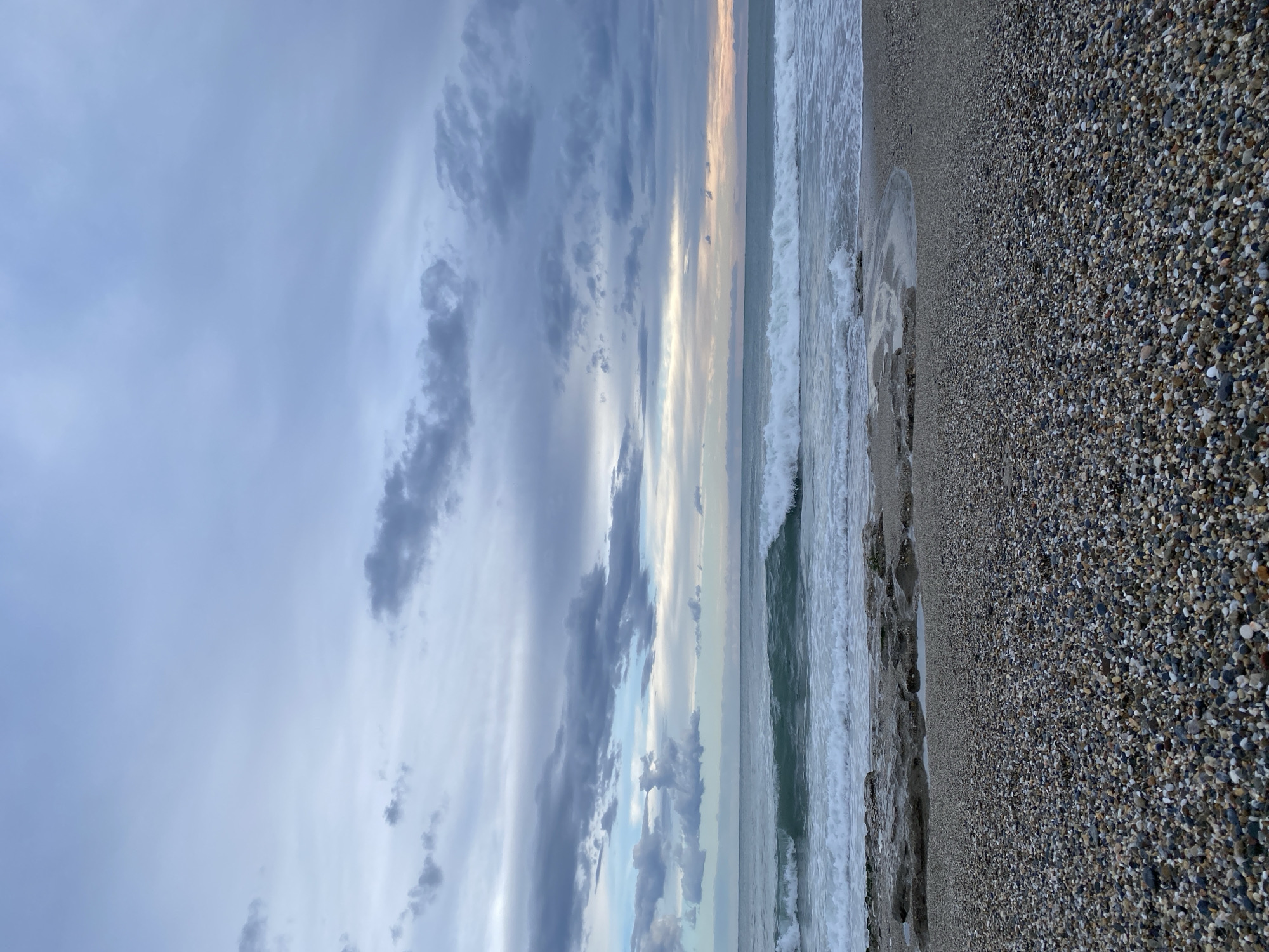 The image captures a beach scene in Alanya, Turkey. The beach is primarily composed of small pebbles and stones. Waves are visible crashing onto the shore. The sky is filled with a variety of clouds, and a thin line of light is visible near the horizon where the sky and sea meet. No people are visible in the frame.