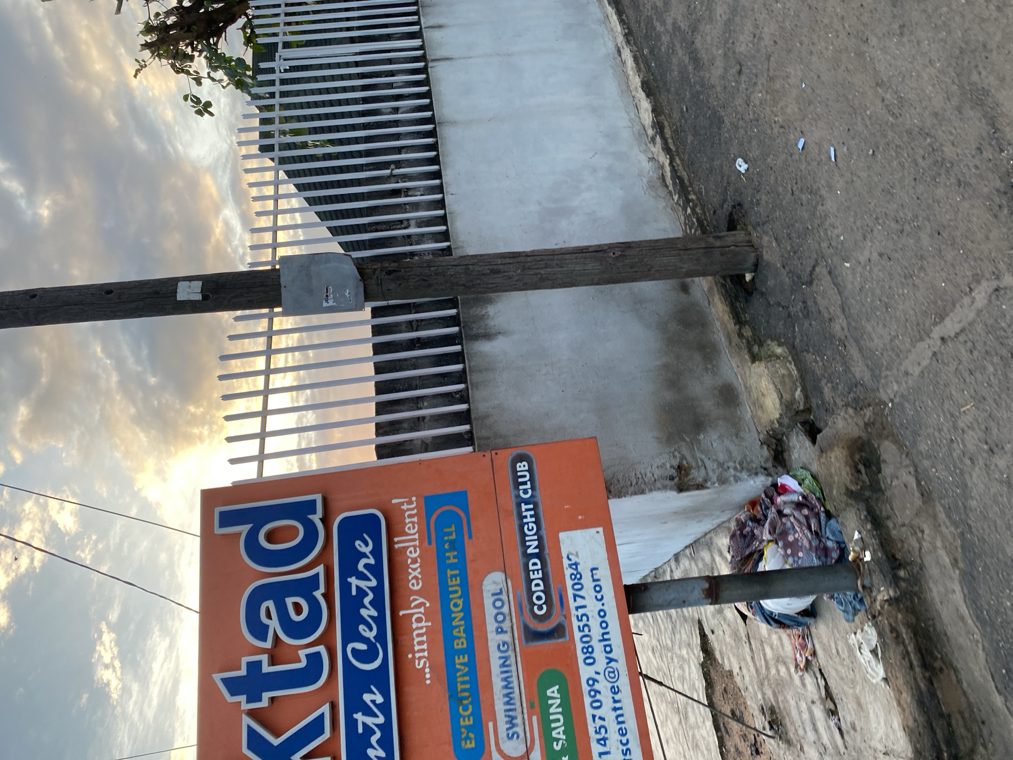 A slightly angled, eye-level shot captures a street-level view of an orange sign advertising "XTAJ" with the subtitle "nits Centre." Below this, the sign lists "EXECUTIVE BANQUET HALL," "SWIMMING POOL," "& SAUNA," and "CODED NIGHT CLUB." Contact information is provided at the bottom of the sign, including a phone number and an email address: "tscentre@yahoo.com." The sign is mounted on a metal pole. To the right of the sign is a wooden utility pole with some attached electrical equipment. Behind the sign and pole is a white vertical slatted fence, and beyond that, a building with a grey concrete wall. The sky above is partly cloudy with a sunset glow. The ground in the foreground is asphalt with debris and a pile of colorful fabric scraps near the base of the metal pole. The overall scene suggests an outdoor location in a commercial or entertainment district, possibly in Abeokuta, Nigeria, given the context of the sign's name.