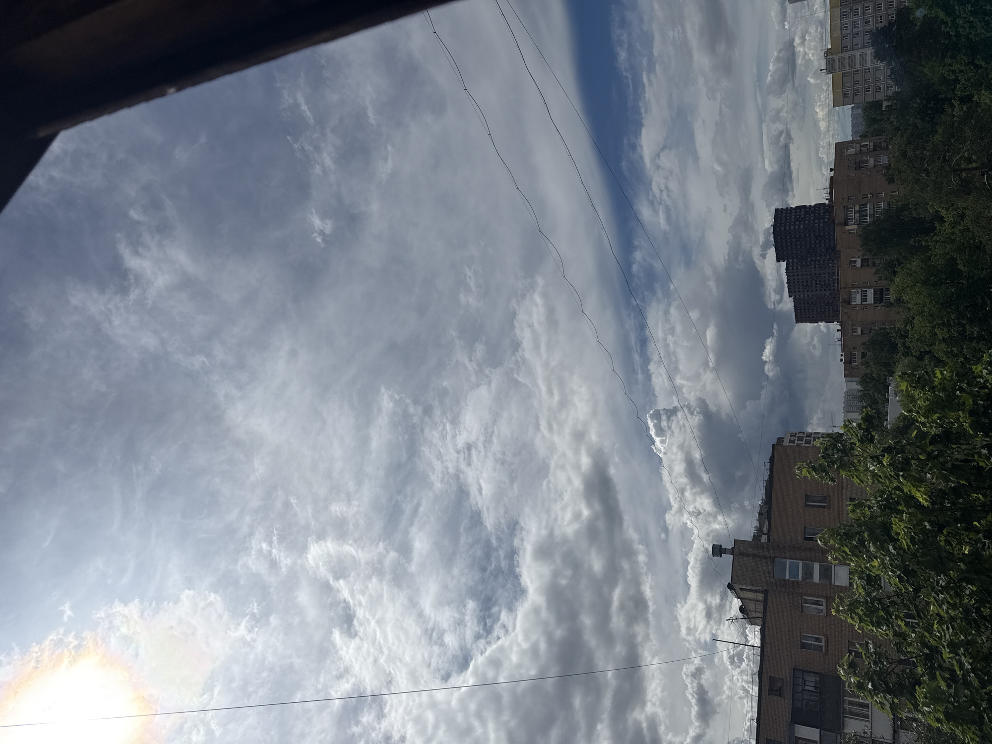 The image shows a view of the sky with clouds, some buildings, trees, and power lines, likely taken from a window or balcony in Mytishchi, Russia. The sky is filled with various cloud formations. The buildings appear to be residential apartments. Green trees are visible on the right side of the image. Several power lines stretch across the sky. The sun is also visible in the lower left corner.