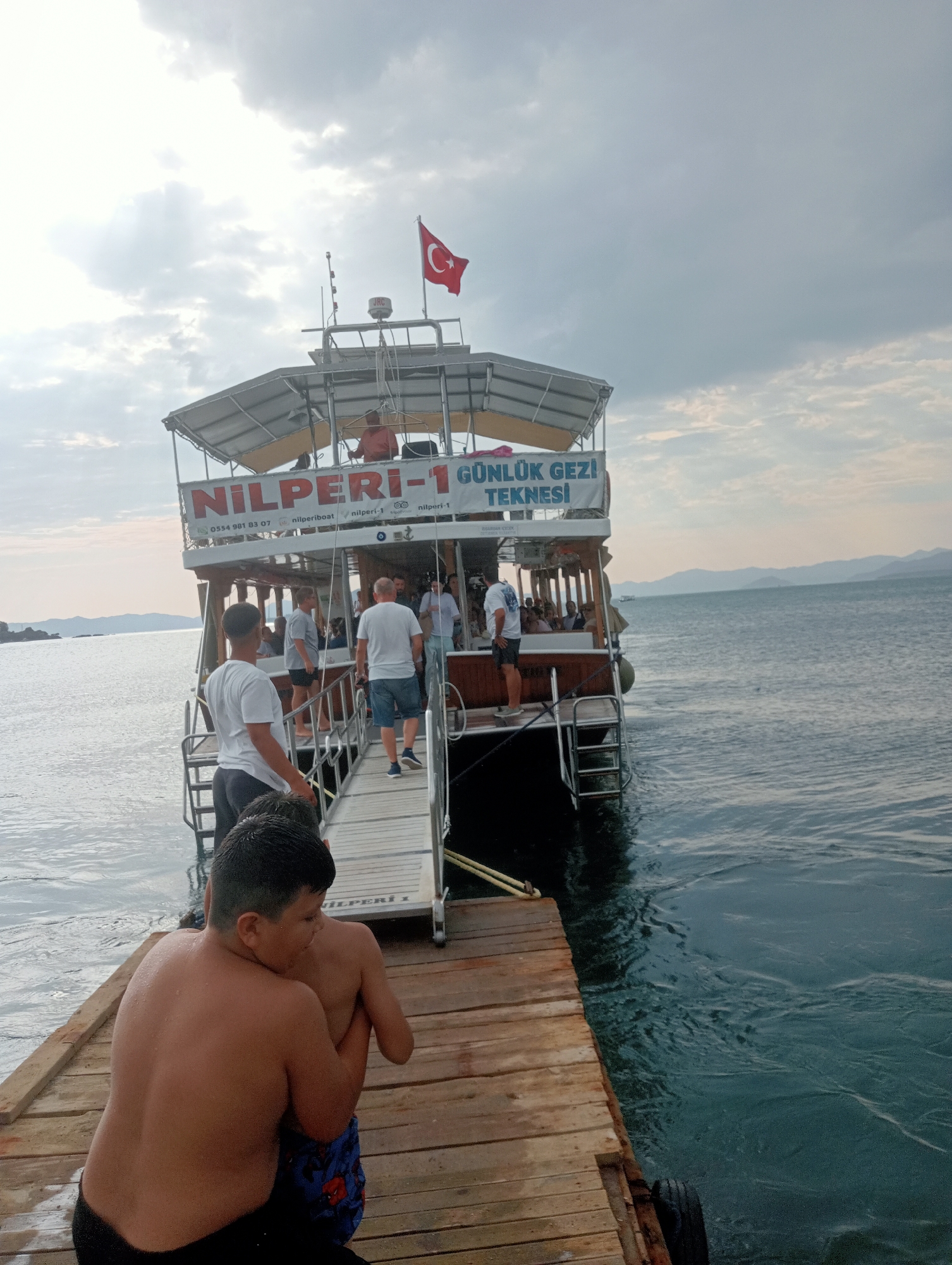 A group of people are boarding a boat from a wooden pier. The boat has "NILPERI-1 GUNLUK GEZI TEKNESI" written on its side, indicating it is a daily tour boat named Nilperi-1. Two young boys are on the pier in the foreground, one embracing the other from behind. The boat is docked on the water, with calm, dark blue water surrounding it. In the background, distant landmasses are visible, and the sky is overcast with some clouds. The scene appears to be a tourist excursion in Fethiye, Turkey.