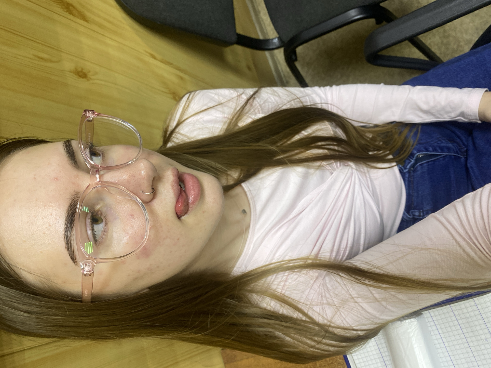 This media file is a portrait of a young woman. She has long, brown hair and is wearing pink-rimmed glasses. Her tongue is sticking out slightly, and she has a nose ring. She is wearing a light pink long-sleeved shirt and blue jeans. The background appears to be indoors, with wooden paneling visible on the left and dark chairs in the background. The lighting suggests it might be daytime, but there are no strong indicators of the time of day or weather. There is no visible text in the image. The overall impression is informal and candid. The subject's expression and pose suggest a playful or casual mood. The setting appears to be a room with simple furnishings, possibly a home or an office.