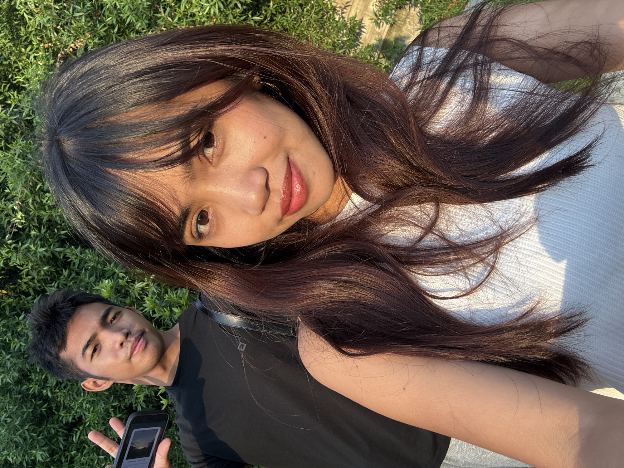 The image shows two individuals, a male and a female, posing for a selfie outdoors with a dense green hedge as the backdrop. The male, positioned in the background on the left, is wearing a black t-shirt and grey shorts. He is holding up a smartphone in his left hand and making a peace sign with his right hand, looking towards the camera with a slight smile. The female is in the foreground on the right, wearing a white ribbed tank top. Her long, dark brown hair frames her face, and she is looking directly at the camera with a subtle smile and a touch of lipstick. The sunlight casts shadows across the scene, indicating it is likely late afternoon. The environment suggests an outdoor setting, possibly a park or garden area within Davao City, Philippines.