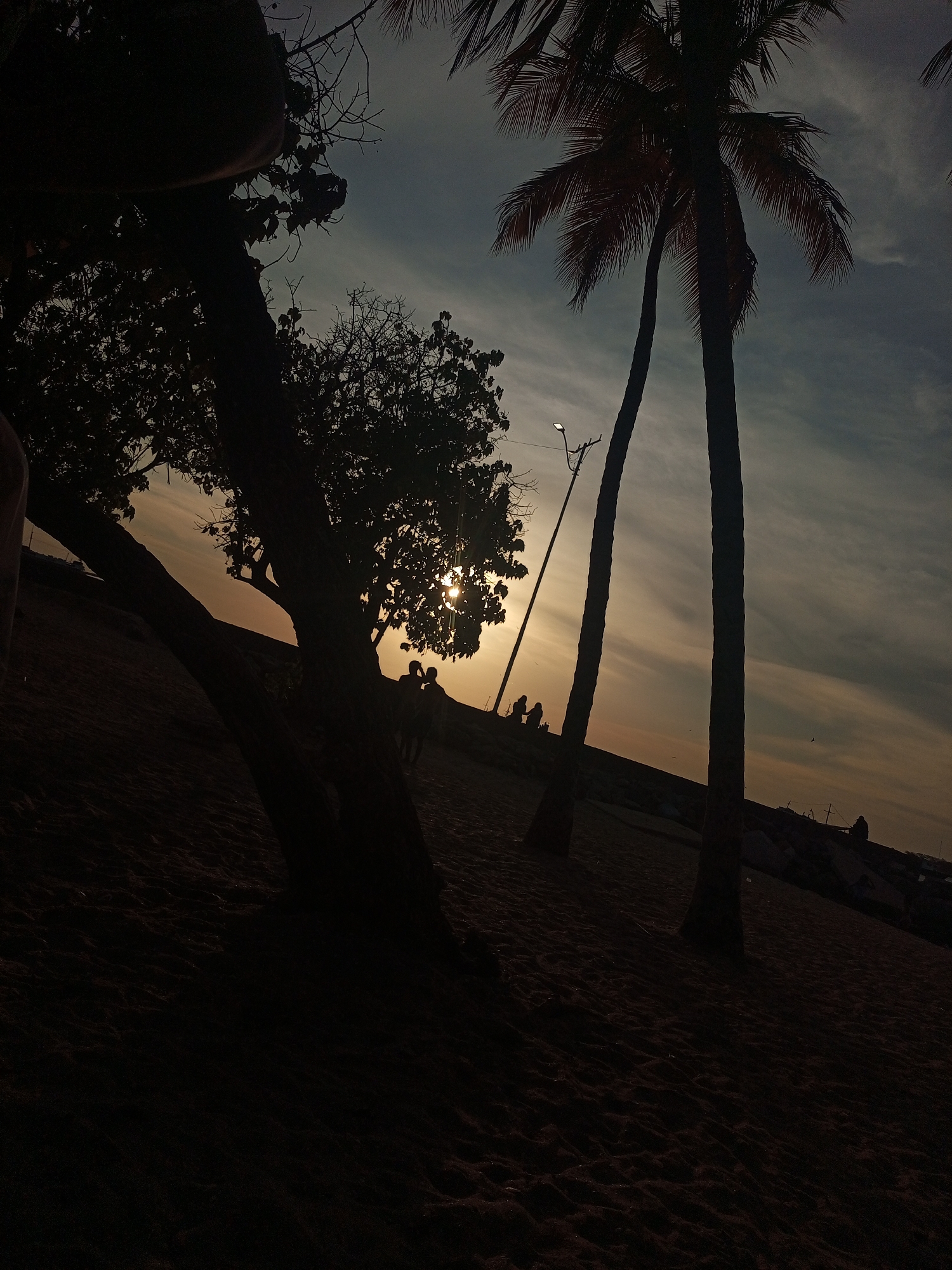 The image shows a beach scene at dusk, likely in Juan Griego, Venezuela, given the request for location specificity. Silhouetted trees, including palm trees, dominate the foreground. The sun is setting, creating a bright glare behind a tree with people standing near it. Further in the background, more people are visible sitting or standing along what appears to be a waterfront edge. A street light is present. The beach sand occupies the lower portion of the frame and is dark due to the low light. The sky shows a mix of light orange near the horizon and darker blue above, with some cloud formations.