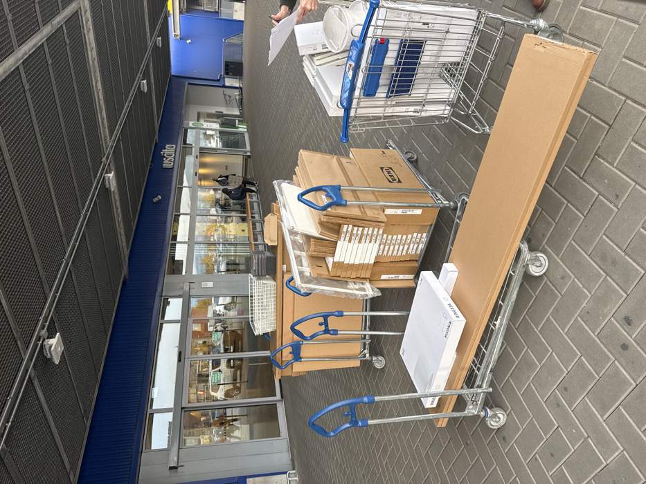 The scene is an outdoor paved area in front of an IKEA store in Sesto Fiorentino, Italy. The building facade features a prominent blue band above automatic glass doors and a dark grey corrugated ceiling. The word "Uscita" (Italian for "Exit") is visible above one set of doors.
In the foreground and midground, four trolleys are loaded with items. A long, flatbed trolley in the immediate foreground carries a large, flat, brown cardboard package and a smaller white box labeled "RINGSTA." Behind it, two similar but shorter flatbed trolleys are stacked with numerous brown cardboard boxes, many bearing the IKEA logo, indicative of flat-pack furniture. One of these trolleys also holds a white plastic basket. To the right, a standard metal shopping cart with blue handles is filled with various white and metallic household items, including a waste bin and possibly a lighting fixture.
On the far right, partially visible, are the lower arms and hands of two individuals; one holds a clipboard with papers. In the background, two individuals are seated on a wooden bench located in front of the store's glass entrance/exit. Reflections in the glass doors show a white Jeep and other indistinct figures in a parking area beyond. The paving is composed of grey bricks arranged in a herringbone pattern.