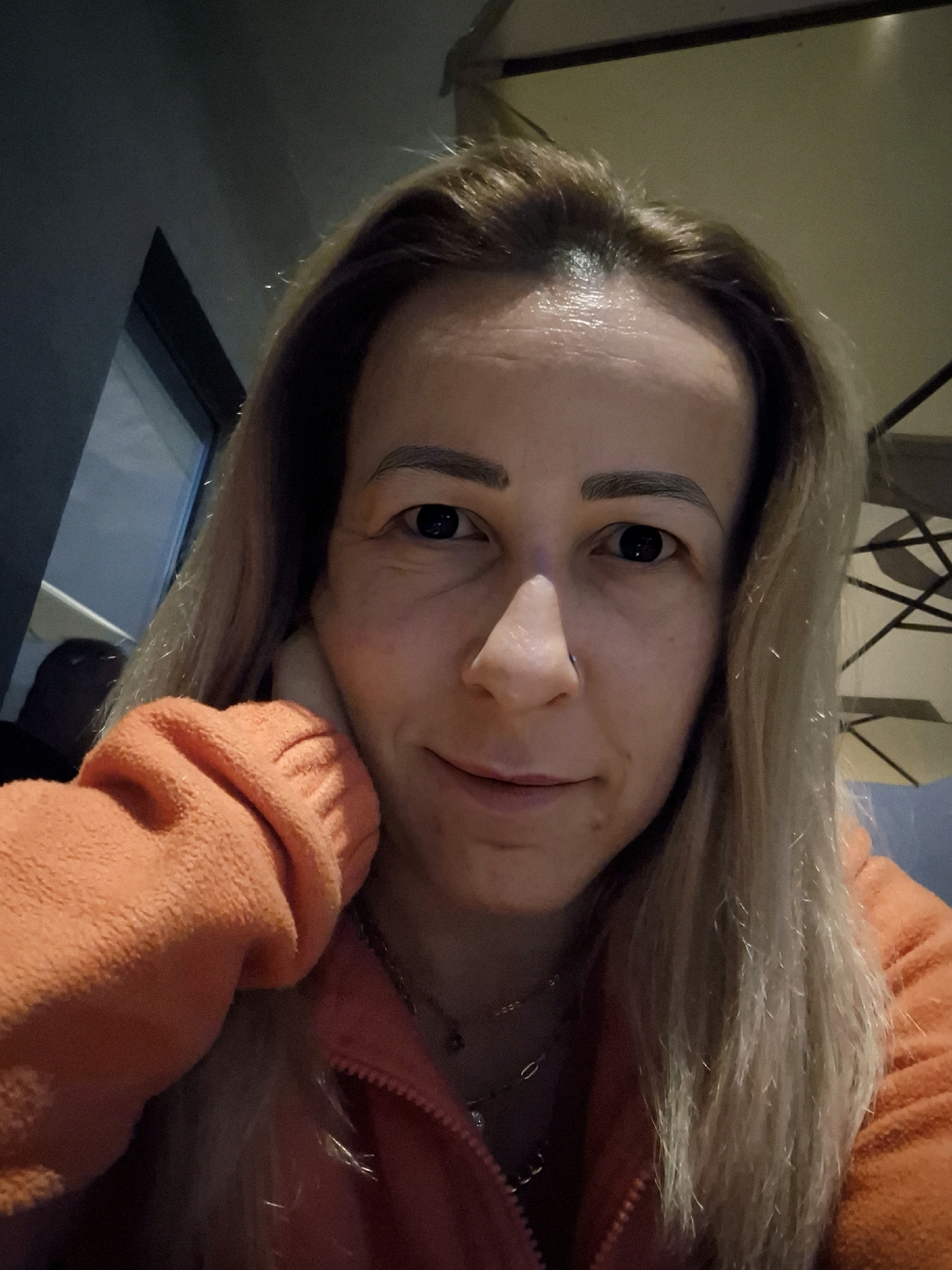 A medium shot captures a young Caucasian woman with blonde hair and light skin, resting her right hand on her cheek and looking directly at the camera. She is wearing an orange hooded jacket. The background is dimly lit, with indistinct shapes of furniture and possibly a window suggesting an indoor setting. The lighting is low, casting shadows and highlighting the contours of her face.