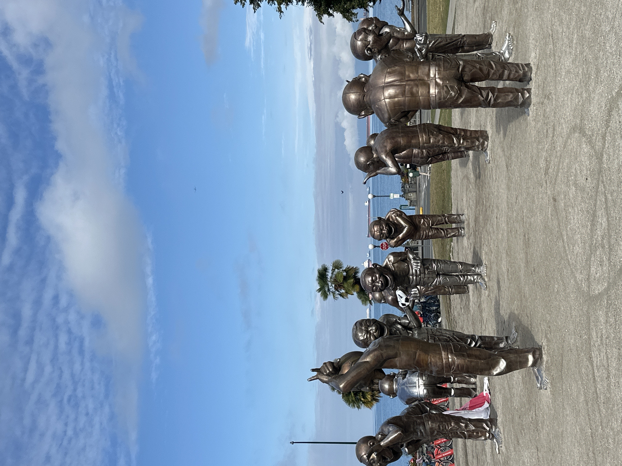The image shows the "Laughing Man" sculpture series by artist Yue Minjun, located on the waterfront in Vancouver, Canada. The bronze sculptures depict figures with exaggerated, laughing expressions and are arranged in a circle on a paved surface. In the background, the ocean is visible, with several ships in the distance, a palm tree, a street lamp, and a stop sign. The sky is blue with scattered clouds, and a faint outline of a distant mountain range can be seen. The overall scene conveys a sense of public art in an outdoor urban environment.