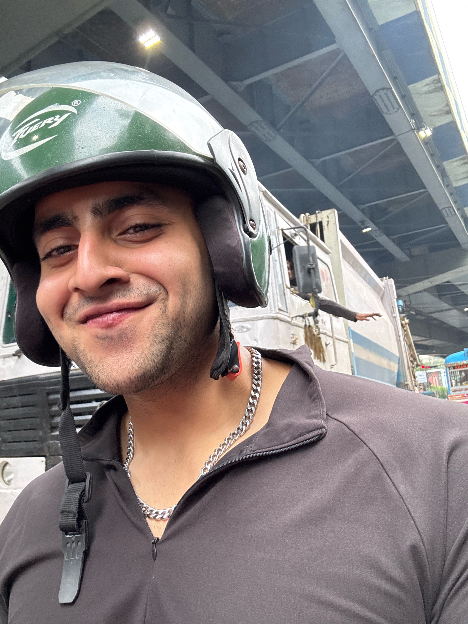 The image shows a close-up selfie of a man wearing a green and black helmet and a black zipped jacket with a silver chain. The man is smiling and looking at the camera. In the background, a large white garbage truck is visible, with a person leaning out of the driver's side window with their arm extended. The scene appears to be on a city street, possibly under an overpass. The presence of a garbage truck and the overall urban setting suggest a location in Kolkata, India. The lighting indicates it is daytime.