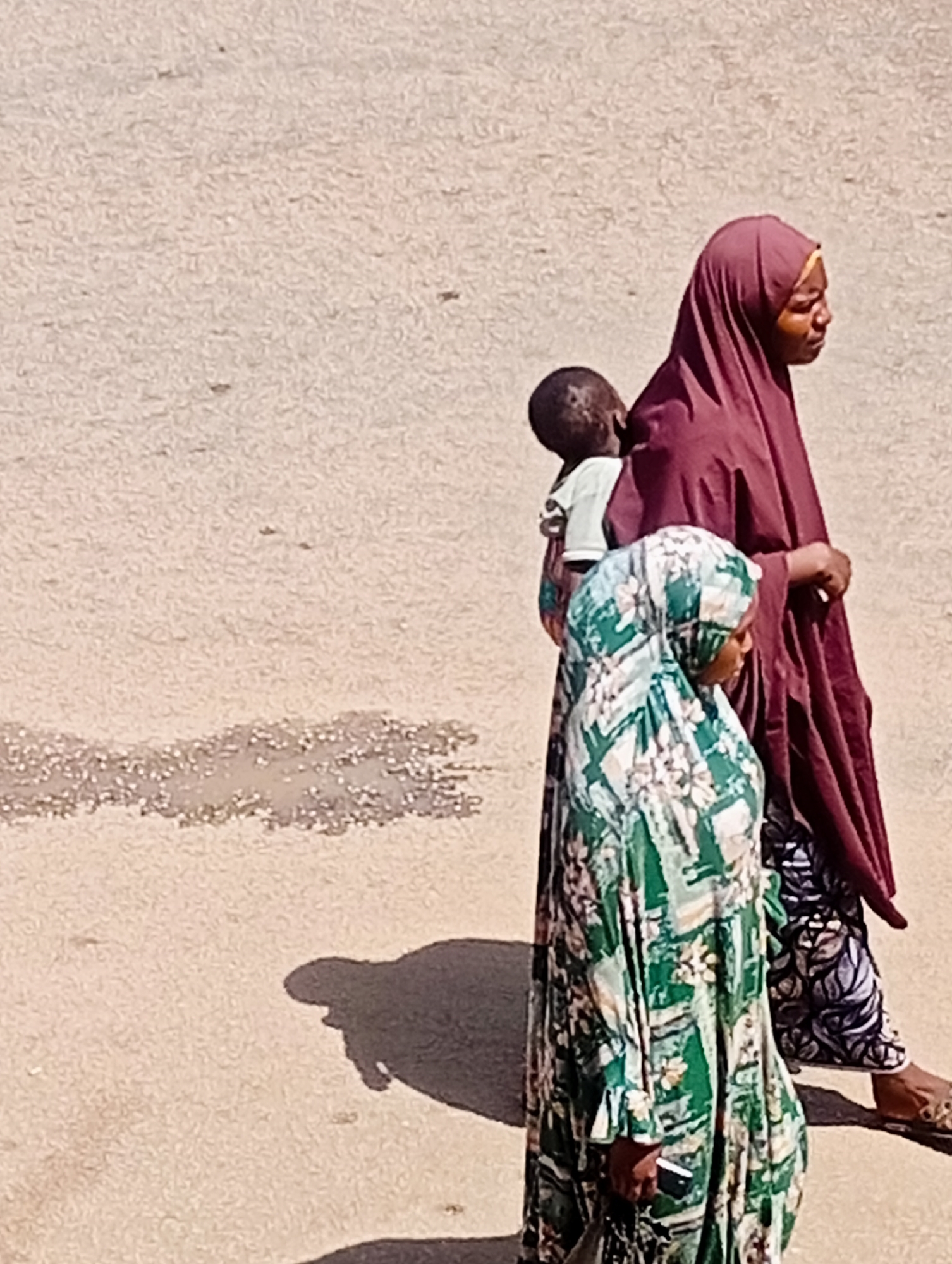 In Bukuru, Nigeria, a group of three people walks on a textured, light-colored dirt or asphalt surface. The foremost individual, on the right, wears a deep red hijab and is carrying a small child on their back, whose head is visible. To the left of this pair, another person walks, wearing a green and white patterned dress and a matching hijab. Their heads are both turned to the right, suggesting they are looking in the same direction. The ground in the foreground shows some damp or discolored patches and a shadow cast by the individuals. The overall lighting suggests it is daytime, with the sun casting sharp shadows.