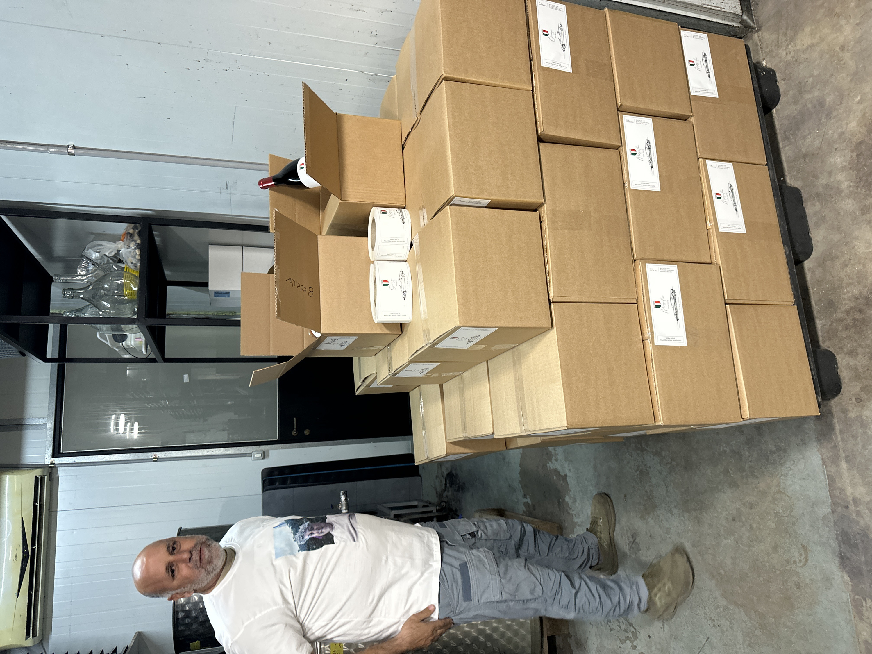 An interior view of a commercial or industrial facility in Tira, Israel, depicts a bald, bearded adult male standing next to a large stack of cardboard boxes. The man, positioned on the left side of the frame, is wearing a white t-shirt with a graphic print, grey cargo pants, and olive-green sneakers; his right hand is in his pocket, and he is looking directly towards the camera.
In the mid-ground, a substantial quantity of unsealed and sealed brown cardboard boxes are stacked on a black plastic pallet. Two boxes on the top layer are open, with one revealing a dark-colored bottle with a red capsule, indicative of a wine bottle. Multiple boxes feature white labels bearing an Italian flag emblem, the word "Orange" or "Maserati" in script, and a stylized drawing of a vintage sports car. Two rolls of identical labels are resting on a box in the upper right portion of the stacked merchandise.
The background features light-colored paneled walls with vertical seams. An off-white Midea air conditioning unit is mounted high on the left wall. To the right of the man, a dark, cylindrical metallic tank is partially visible. A black metal shelving unit against the back wall holds several clear glass carboys and miscellaneous items, including plastic bags. A dark-colored door or entryway is visible in the left background. The floor is concrete with visible discoloration and stains.