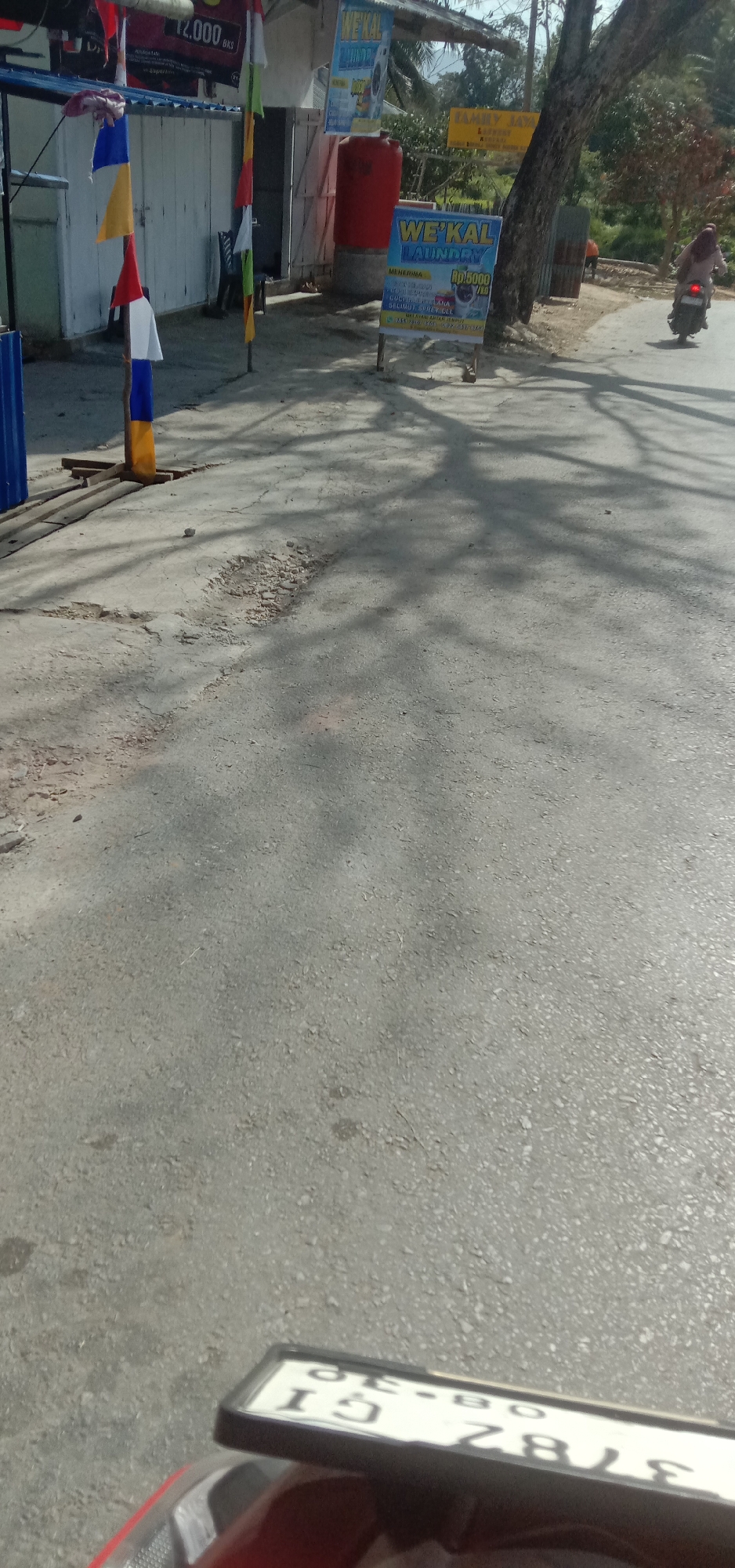 The image depicts a street scene in Kendari, Indonesia, characterized by a slightly weathered asphalt road. The road surface shows signs of wear and tear, with some patches of exposed aggregate and minor cracks. The lighting suggests it is daytime, with shadows cast by trees and structures.
In the background, a building with white-paneled doors and a corrugated metal roof is visible. Flags in various colors, including red, white, blue, and yellow, are displayed on poles in front of the building, indicating a festive or local event. A large red water tank stands next to the building.
A prominent sign, "WE'KAL LAUNDRY," is displayed on a blue easel. The sign advertises laundry services with pricing information: "MENERIMA... RP5000 /KG". Another yellow sign, partially visible, reads "FAMILY JAYA LAUNDRY".
Further down the road, a person is riding a motorcycle. The motorcycle is dark-colored, and the rider is wearing a light-colored headscarf and clothing. The motorcycle's license plate is visible in the foreground, displaying characters that appear to be "GI 378Z BIK".
The overall environment suggests a commercial or residential area within Kendari, with elements of local businesses and community presence.