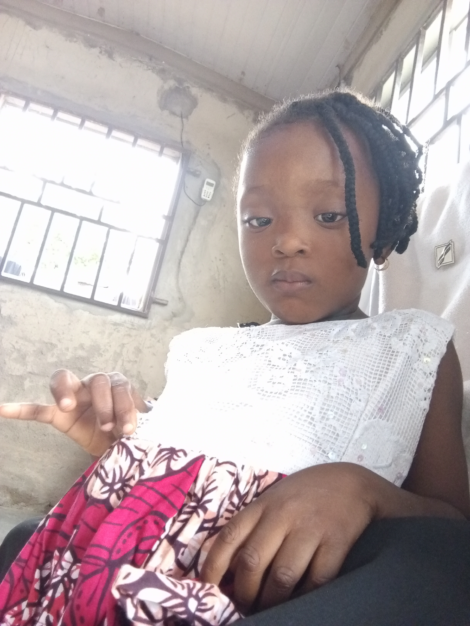 This is a portrait of a young Black girl, likely a child, seated indoors. She has dark skin and braided hair, with one braid falling forward over her forehead. She is wearing a white lacy top over a patterned fabric, which appears to be predominantly pink, black, and white with floral motifs. Her left hand is raised, with her index finger extended, as if pointing or making a gesture. Her right arm is bent, and her hand rests on her lap.
The background suggests an indoor setting with a somewhat unfinished or rustic aesthetic. To the left of the girl is a window with metal bars, through which bright daylight is visible, indicating it is daytime. The walls appear to be concrete or a similar rough material. A white electronic device, possibly a thermostat or a small digital display, is mounted on the wall near the window, with a black wire connected to it. The lighting in the scene is bright, suggesting a sunny day. The girl's expression is neutral, with her gaze directed towards the camera. The overall impression is of a candid, perhaps informal, photograph taken within a home or a similar residential environment in Lokoja, Nigeria.