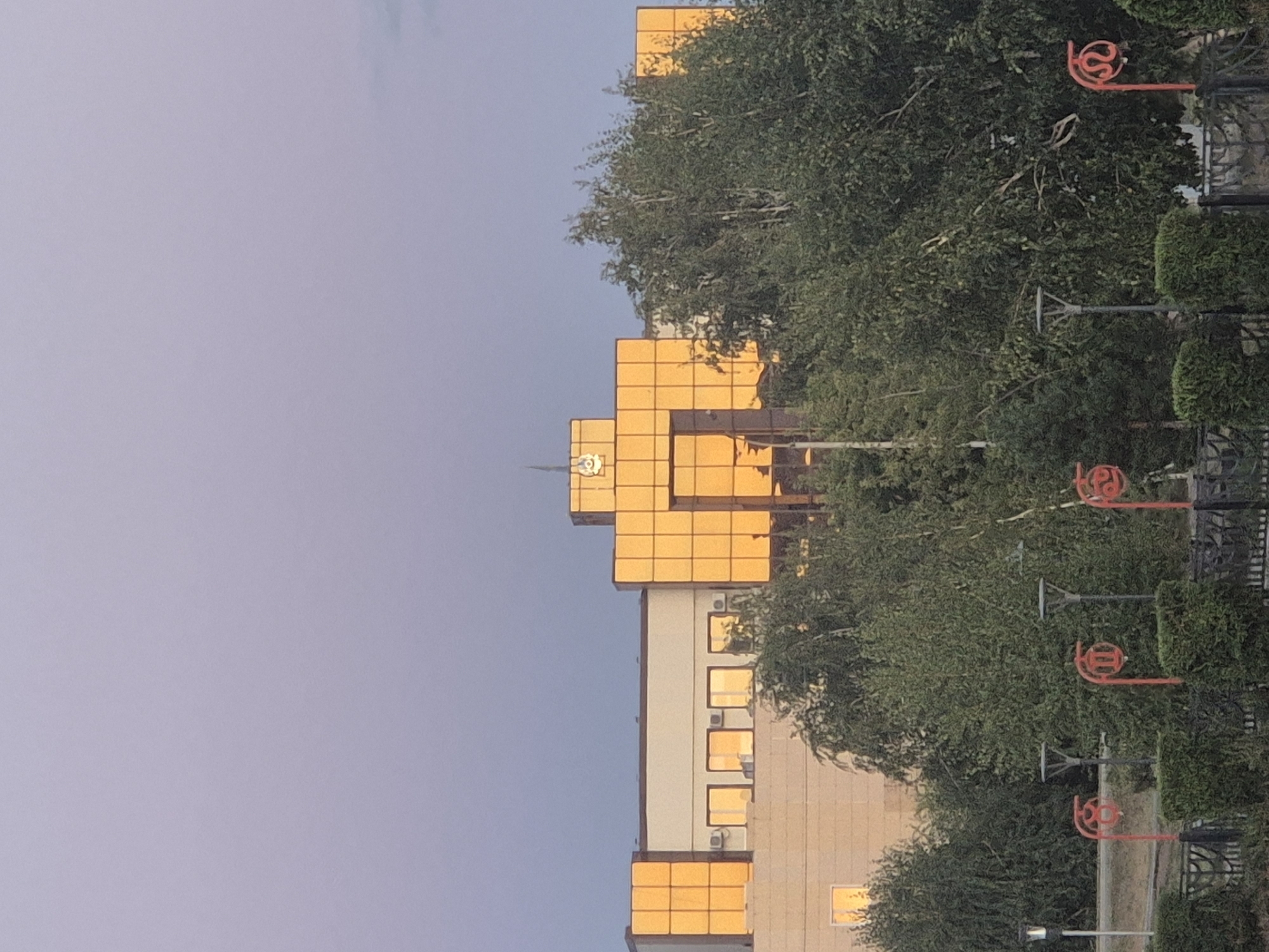 The image shows a building with a gold-colored facade and large windows reflecting the sunset, located in Kokshetau, Kazakhstan. The building appears to be an administrative or commercial structure. In the foreground, there is lush greenery with trees and bushes, and a black metal fence. Several decorative red lampposts with circular designs are visible along the walkway. The sky is a soft gradient of purple and grey, suggesting it is dusk or dawn. There are no people or vehicles visible in the image.