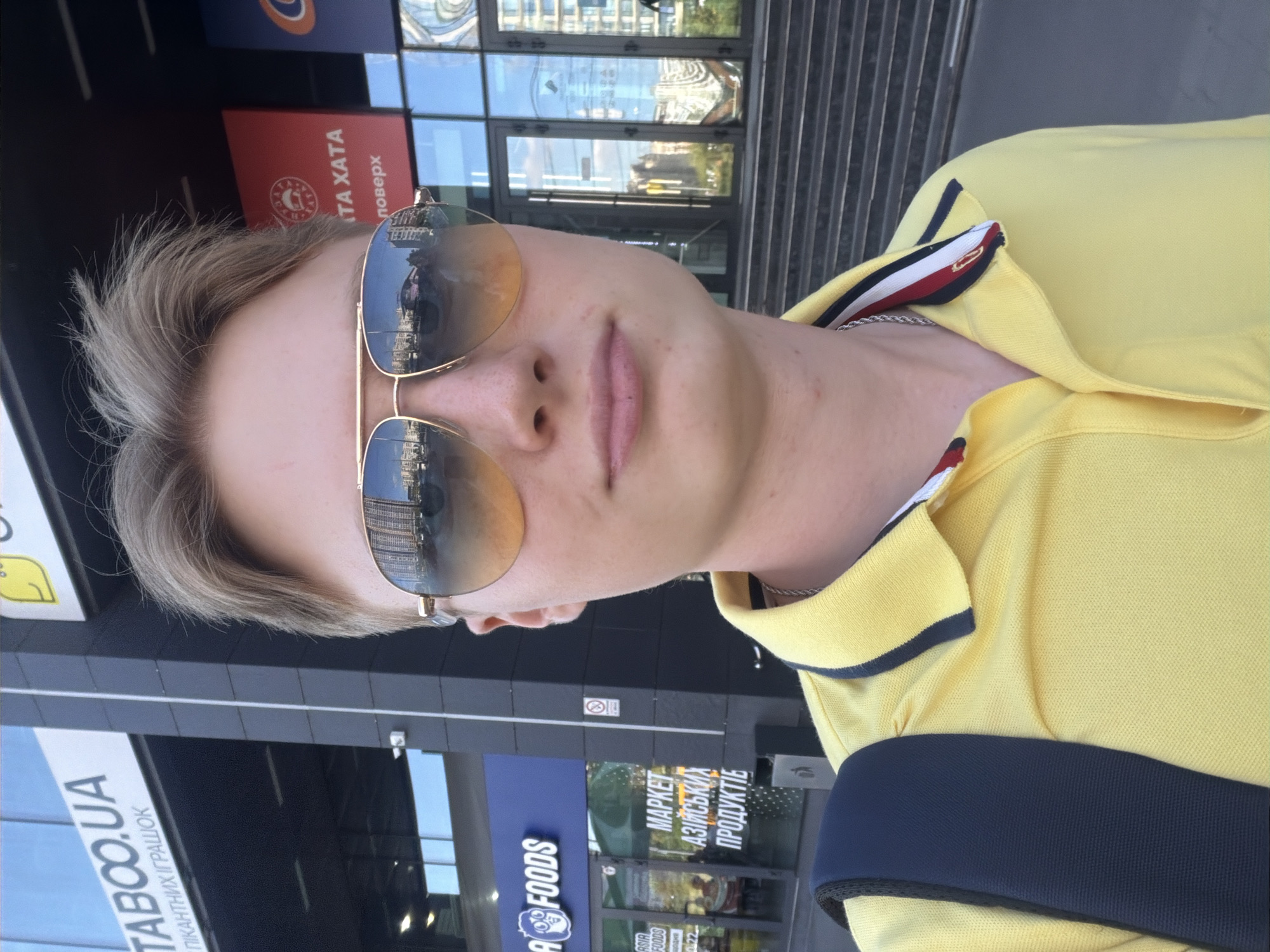 A young man wearing aviator sunglasses and a yellow polo shirt stands in front of a modern building with a glass facade. The sunglasses reflect a cityscape, showing buildings and a street. The building has signs that appear to be in Ukrainian, including "TABOO.UA" and "ASIA FOODS." The man is looking towards the camera, and a dark backpack is visible over his left shoulder. The environment suggests an urban setting in Kyiv, Ukraine.