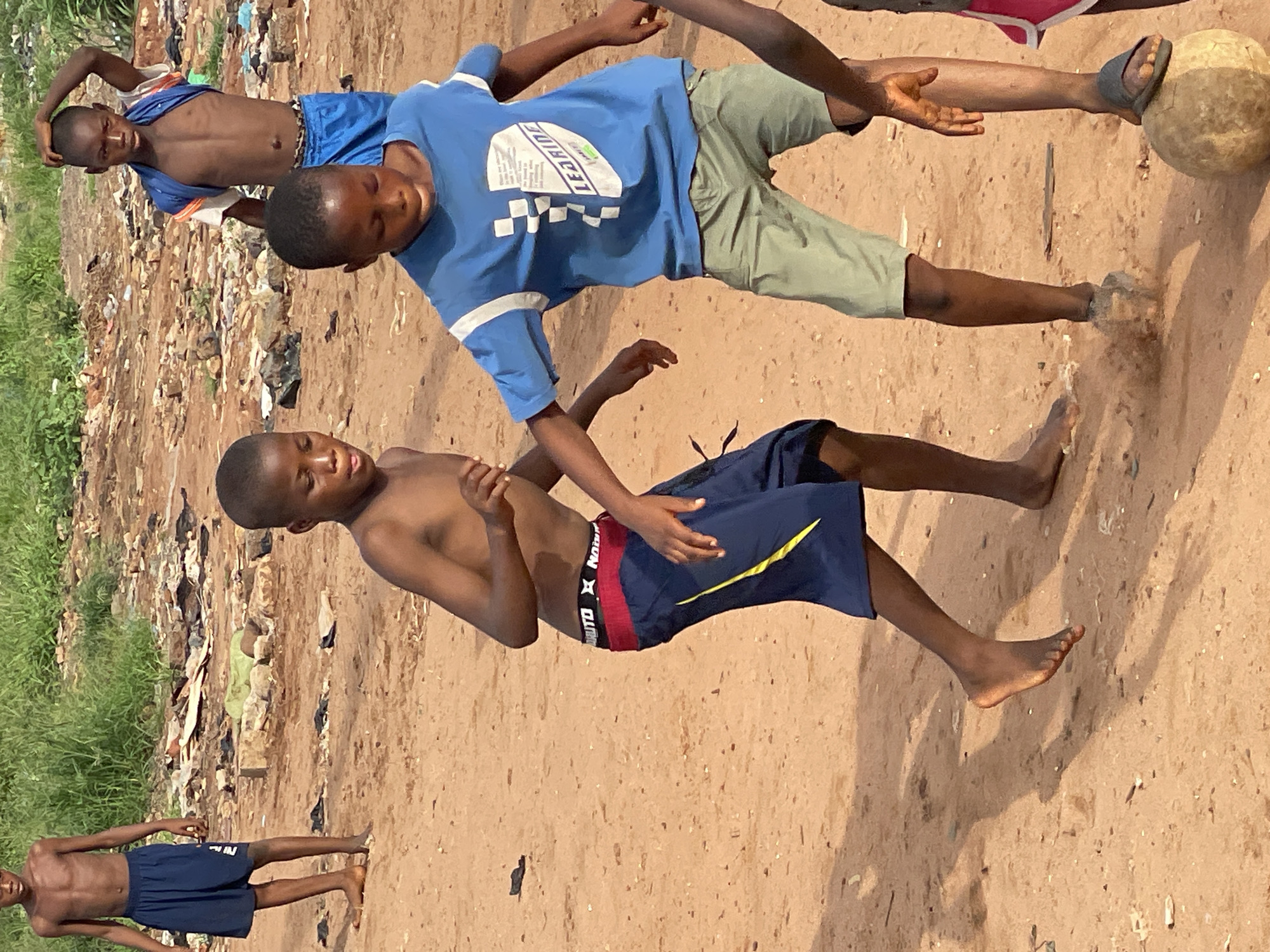 The scene depicts four shirtless boys on a dirt ground playing with a worn soccer ball. One boy wears a blue t-shirt and khaki shorts. Another boy is wearing dark blue shorts with yellow stripes and red detailing at the waistband. Another boy wears a blue outfit. In the background there is trash and green foliage. The location is assumed to be Onitsha, Nigeria.