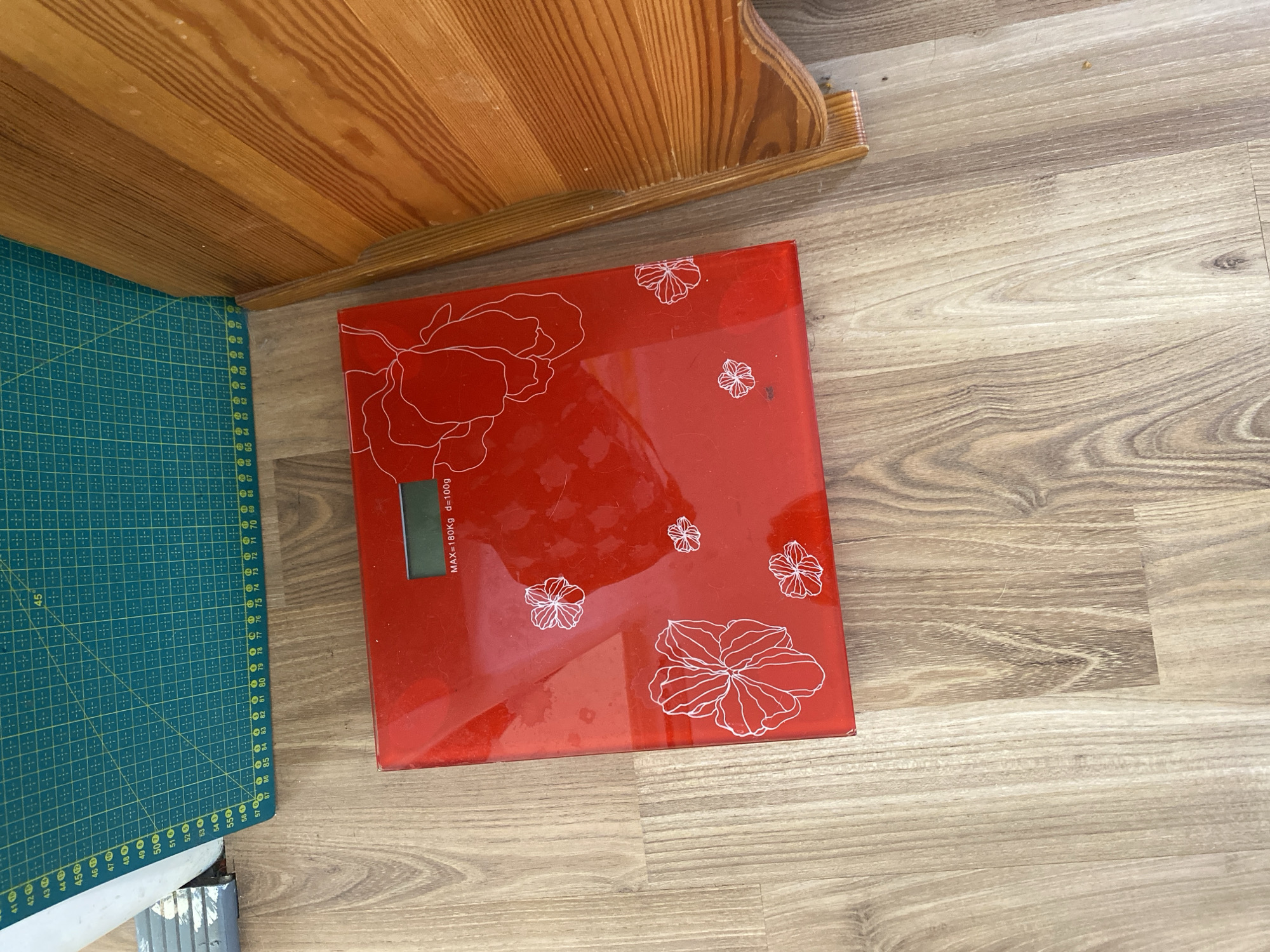 A red digital scale with a floral design is placed on a wooden floor, next to a green cutting mat. The scale has a digital display that reads "MAX=180Kg d=100g". The cutting mat has grid lines and markings that appear to be measurements. A wooden chair or furniture piece is visible on the right side of the image. The scene appears to be indoors, likely in a residential setting in Bílina, Czechia, though this is not explicitly stated in the image.