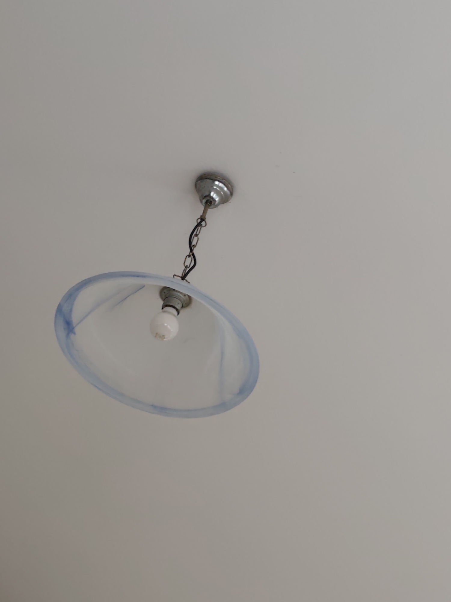 This media file shows a close-up, low-angle shot of a hanging light fixture against a plain, light-colored ceiling. The fixture consists of a chrome-colored ceiling mount, from which a black chain suspends a light blue and white glass lampshade. Inside the lampshade, a single, white, light bulb is visible. The lampshade has a somewhat oval or bowl shape, with a translucent quality and subtle swirling patterns of blue marbling. The overall scene is simple and domestic, with no people or significant activity taking place. The lighting suggests it could be daytime, as the ceiling is brightly illuminated, but the artificial light from the bulb is also present. There are no visible text elements in the image. The setting appears to be an indoor environment, possibly a home or a simple commercial space.