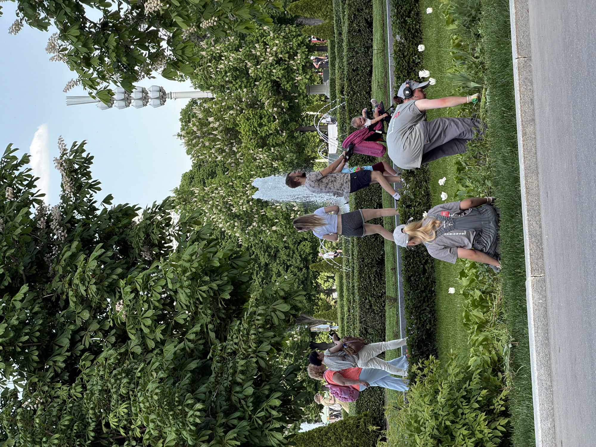 The image shows a park scene in Moscow, Russia. In the background, there is a fountain, trees with white flowers, and a unique street lamp. Several people are present: one man is holding a child in a pink stroller near the fountain, two women are kneeling and tending to the grass and flowers, and other people are walking in the background. A paved pathway borders the green space. The environment is lush with greenery and well-maintained.