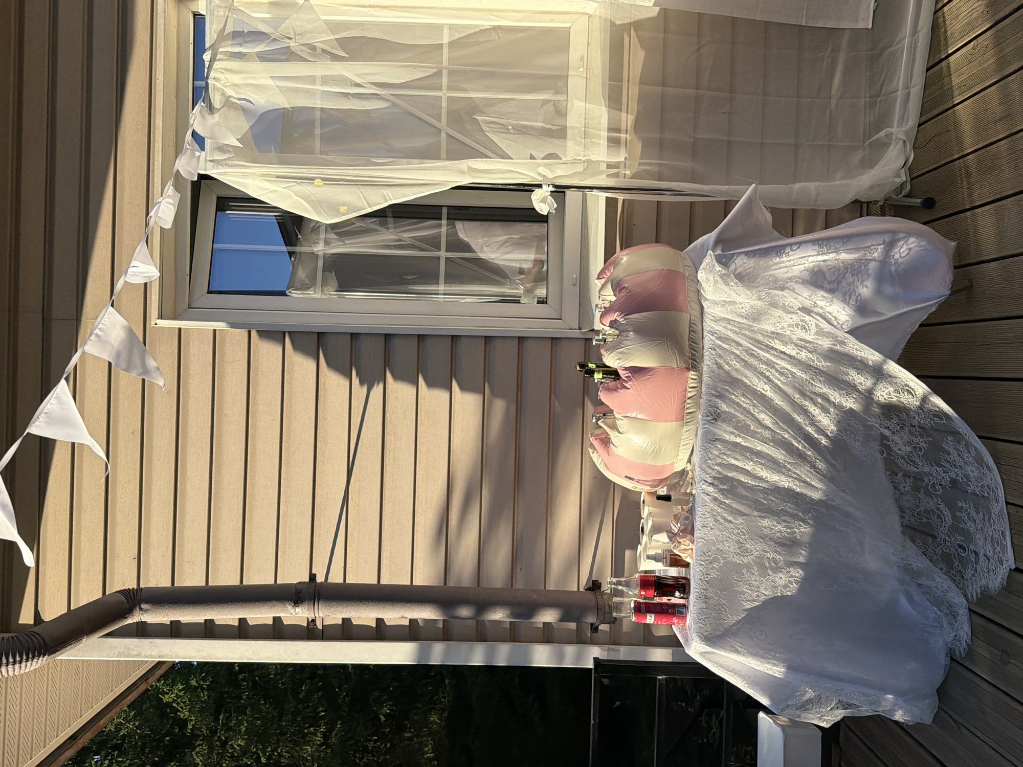 The image depicts a table set up outdoors, possibly for a celebration, on a wooden deck in front of a house with beige siding. A downspout runs vertically along the side of the house. A string of white triangular bunting is strung across the top of the frame, partially obscuring the upper portion of a window and a sheer white curtain hanging to the right of the window.
On the table, which is covered with a white lace tablecloth, are several items. These include two red cans, a bottle of Coca-Cola, several small white cups, a small bottle of liquid, and a pink and white striped inflatable object, possibly a decorative element or a cooler. The inflatable object is placed centrally on the table. Sunlight casts long shadows across the scene, indicating it is late afternoon or early evening. The surrounding environment includes trees visible on the left side of the image. There are no people present in the image. The location is identified as Novosaratovka, Russia.