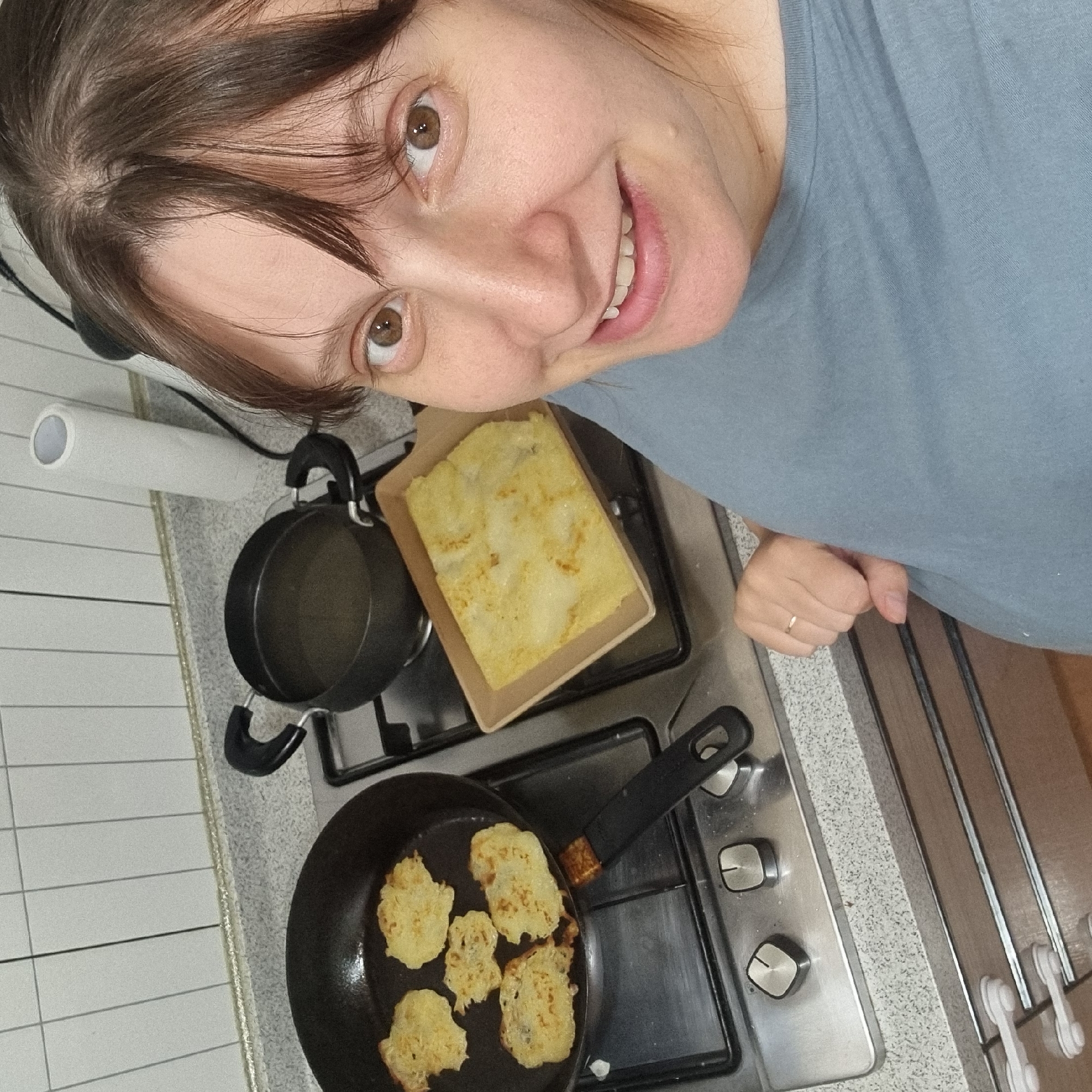 In a kitchen setting, a person with brown hair is smiling and looking at the camera. They are wearing a light blue t-shirt. In the background, a stovetop is visible with a frying pan containing several golden-brown, round items, likely pancakes or fritters, and a pot. There is also a square cardboard container with what appears to be a cooked dish, possibly a casserole or baked good, situated on the stovetop. The scene suggests food preparation or cooking. A roll of paper towels is mounted on the wall to the left. The overall lighting indicates an indoor environment, possibly during the daytime.