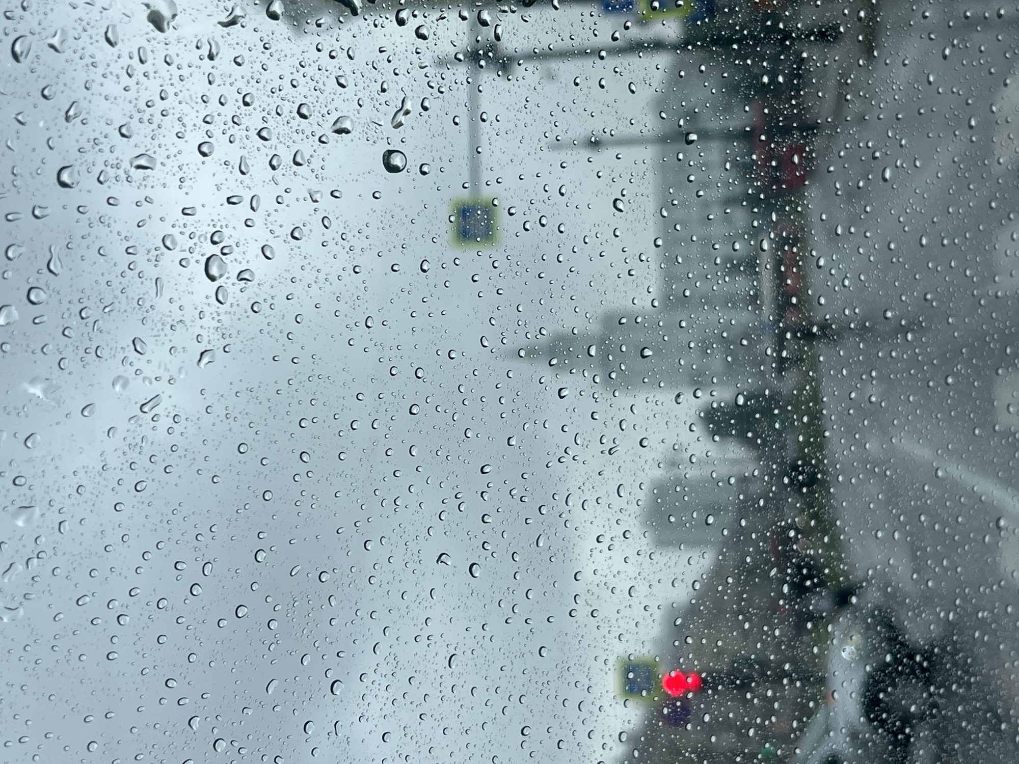 This is a close-up, medium shot of a car windshield covered in water droplets, looking out onto a city street on a cloudy, rainy day. The droplets distort the view of the urban environment behind the glass, creating a blurred, impressionistic scene.
In the background, a traffic light is visible, displaying a red light. Further back, there are blurred buildings, including what appears to be a taller structure with a pointed roof, possibly a spire or tower. A pedestrian crossing sign is also discernible, with a blue square sign. Various vehicles, including cars and buses, are visible as indistinct shapes and colors, such as a red bus. The road surface is wet and reflective, indicating the ongoing rain. Trees and greenery are also present but heavily obscured by the rain and distance.
While the specific location within the city of Perm, Russia, cannot be definitively identified from the visual information alone, the presence of typical urban infrastructure like traffic lights, multi-story buildings, and roads suggests a city environment consistent with Perm. The overall impression is one of a typical rainy day in a Russian city, experienced from inside a vehicle. There are no clearly discernible individuals or specific interactions. The focus is on the atmospheric conditions and the distorted view of the cityscape.