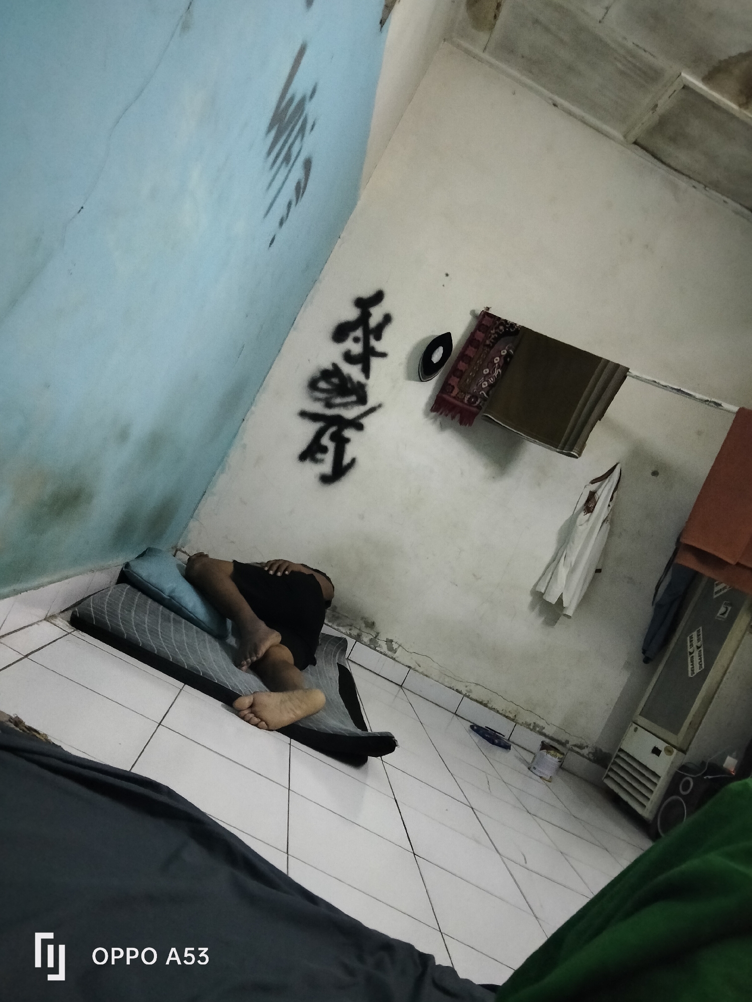 The image depicts a dimly lit room with a tiled floor, likely in Denpasar, Indonesia, given the "OPPO A53" watermark suggesting a locally sold device. The walls are painted a pale blue and white, with visible wear and tear, including cracks and water stains. Black graffiti, possibly in an Asian script, adorns the upper portions of the walls.
In the foreground, a person is lying on their side on a thin mattress with a blue pillow. They are wearing black shorts and their legs are bare. The person is asleep or resting.
The background reveals a clothesline strung across the wall, from which several items are hanging: a dark patterned fabric, a white garment, and an orange textile. To the right, a beige air conditioning unit is visible, with a circular speaker mounted below it. Various other objects, including a dark blue object on the floor and a white container, are scattered around the room. The overall impression is of a simple, lived-in space.