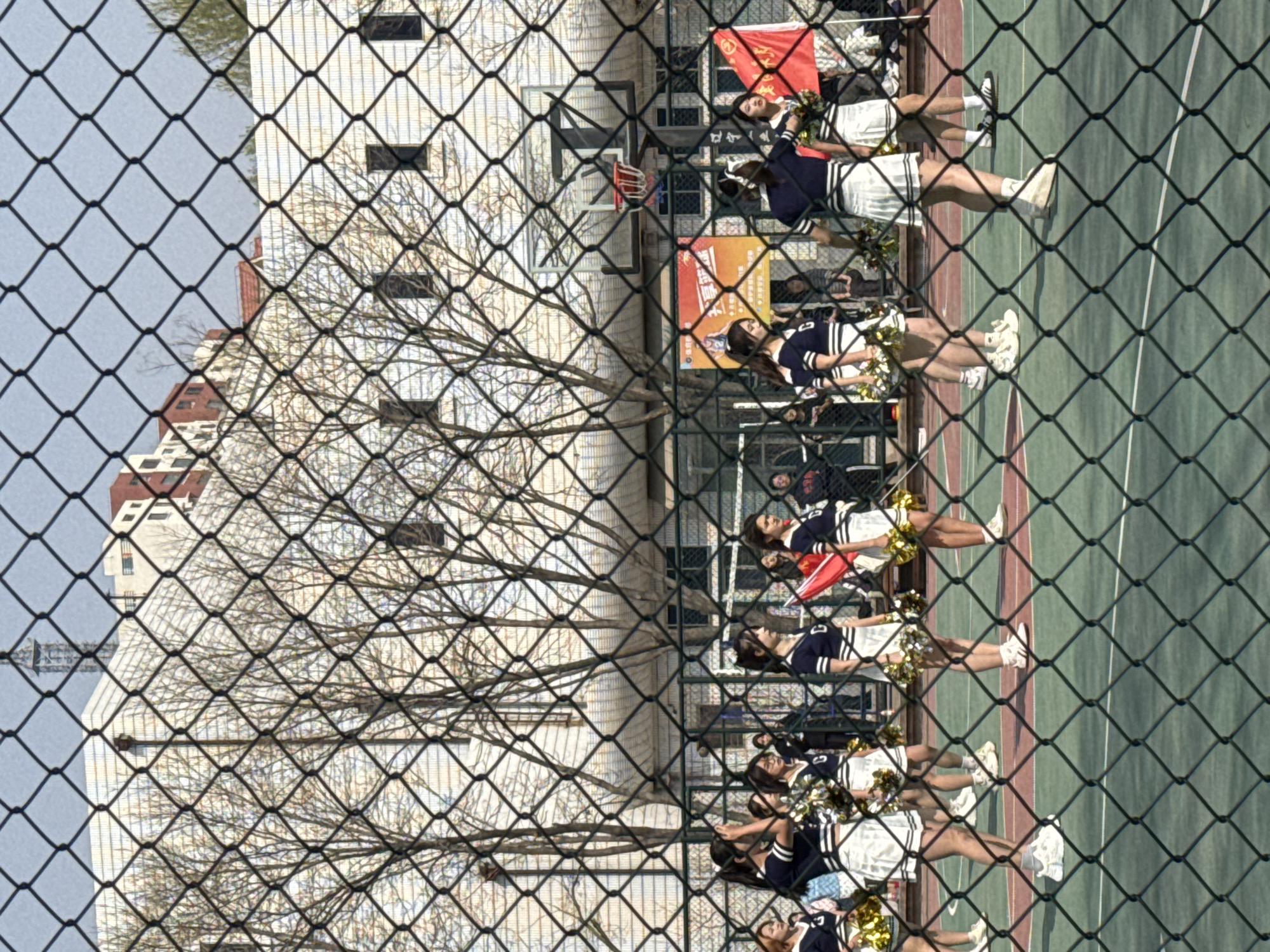 Captured through a chain-link fence, the image depicts a cityscape overlaid with a recreational scene.
In the background, a high-rise building with a grid-like façade and a partially visible red roof is framed by bare tree branches.
Foreground features a group of cheerleaders in matching navy and white uniforms with golden pom-poms, lining the edge of a green and red multi-purpose court. Their faces are visible. A red flag is also seen. Behind the cheerleaders is a fence and some people are standing and sitting behind it.
The vantage point and subjects indicate an outdoor setting, possibly a school or community sports facility.
Given the prompt, the location is inferred to be Zhongtun, China.