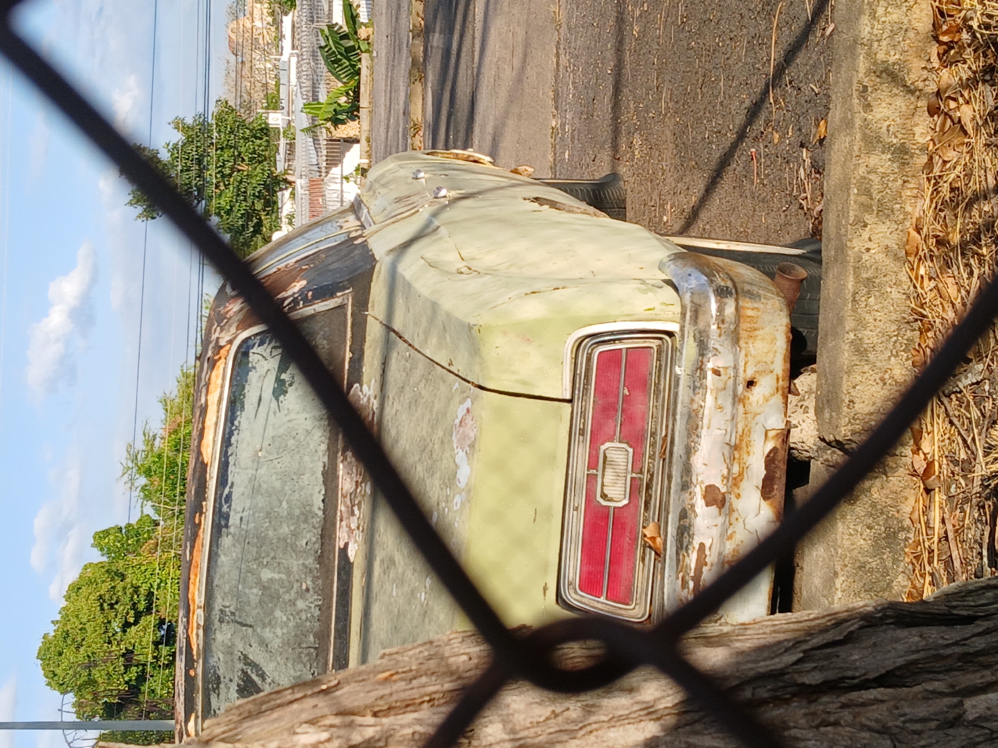 The image depicts an old, dilapidated car parked on a street in Guayana City, Venezuela. The car is visible through a wire fence and is heavily rusted with peeling green paint. The rear of the car is visible, including the tail lights. The car is parked next to a curb and a pile of dry leaves and debris. The street is paved and appears to be in a residential area, with buildings and trees visible in the background. The sky is clear.