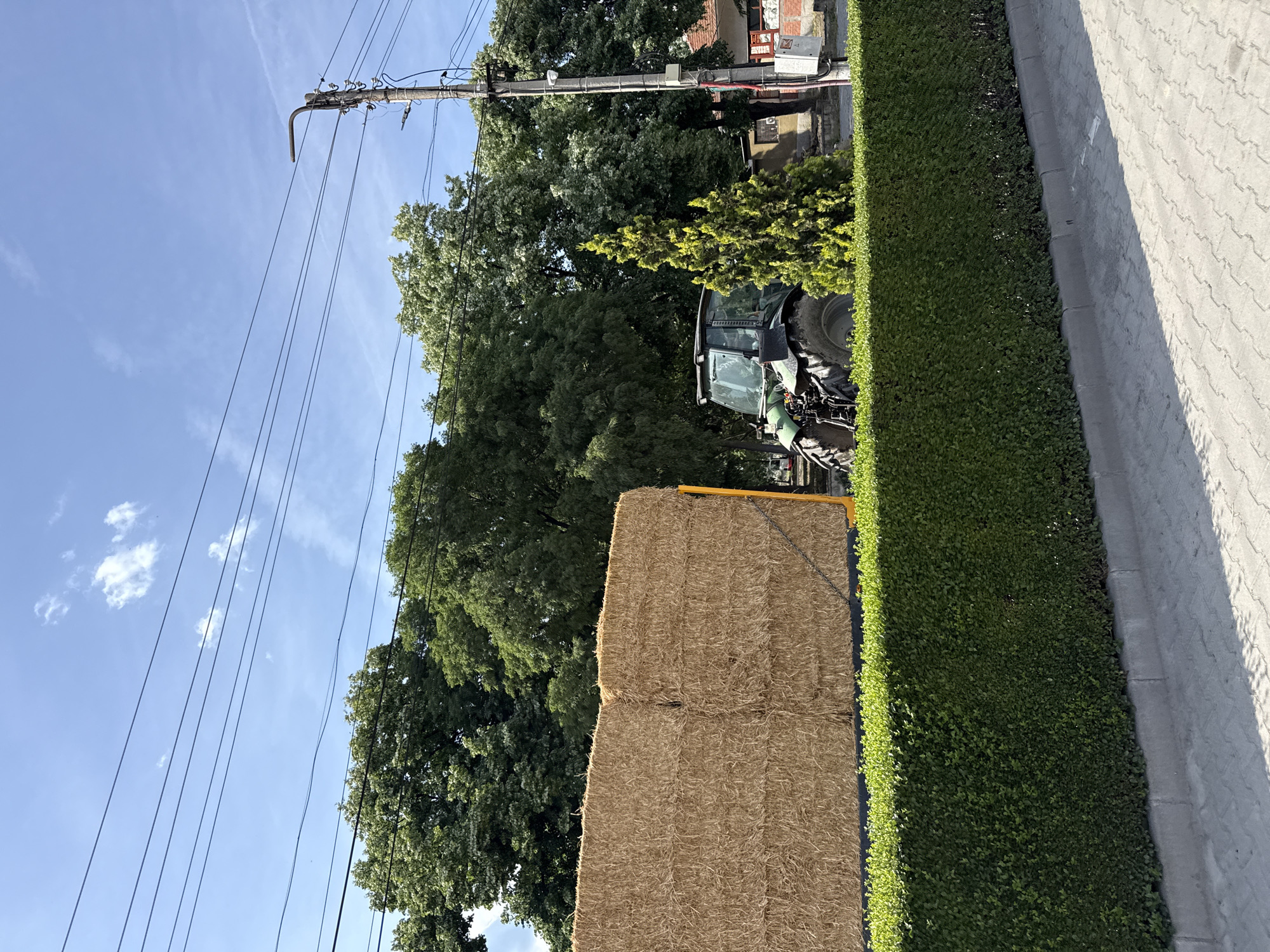 The image shows a scene likely in Katunitsa, Bulgaria, given the presence of Eastern European architectural elements. A large stack of hay bales is prominently displayed near a tall, meticulously trimmed green hedge, suggesting a deliberate arrangement. Adjacent to the hedge is a concrete or paved surface. Beyond the hay bales and hedge, a tractor can be partially seen, hinting at agricultural activity. Utility poles and wires stretch across a blue sky with scattered clouds. In the background, trees are visible, adding to the rural/urban fringe character of the location. A building is further in the background.