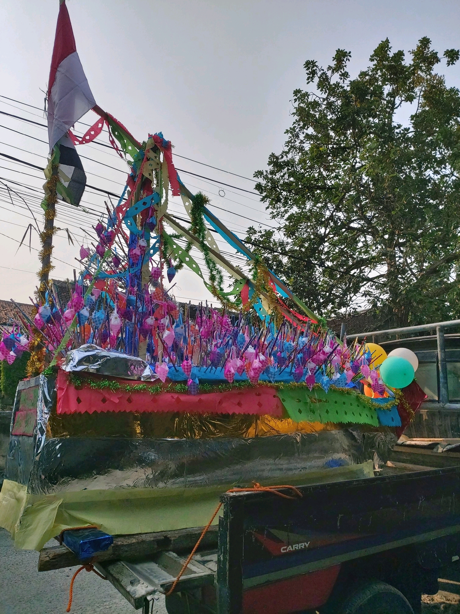 The image shows a decorated float, likely for a parade or celebration, on the back of a truck. The float is designed to resemble a boat and is adorned with numerous colorful paper decorations, streamers, and what appear to be small woven ornaments in various shades of pink and blue. A flag, red and white, is prominently displayed at the top of the float. In the background, there are trees, power lines, and hints of residential buildings, suggesting an outdoor, possibly urban or suburban, setting. Two balloons, one green and one white, are attached to the side of the float. The truck itself is partially visible, with the word "CARRY" visible on its side. The overall scene depicts a festive atmosphere. The location is identified as Bandar Lampung, Indonesia.
