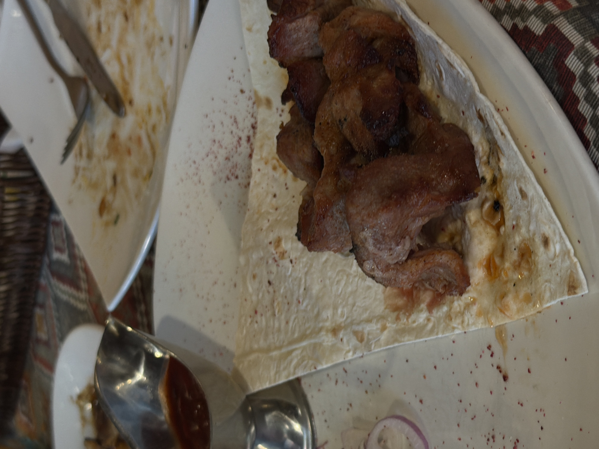 The image depicts a close-up view of a plate of food, likely a kebab or shashlik, served on a piece of flatbread. The meat is grilled and appears to be seasoned. A small metal gravy boat filled with a red sauce is visible to the left of the plate. The plate itself is white and is set on a table covered with a patterned tablecloth. Scattered red spices are present on the plate and the surrounding surface. A few slices of red onion are also visible on the left side of the plate. In the background, other plates and cutlery suggest a dining setting, possibly a restaurant. The overall impression is one of a meal in progress or recently consumed. The image does not contain any discernible landmarks or contextual clues that would allow for specific identification of its location within Saint Petersburg, Russia.